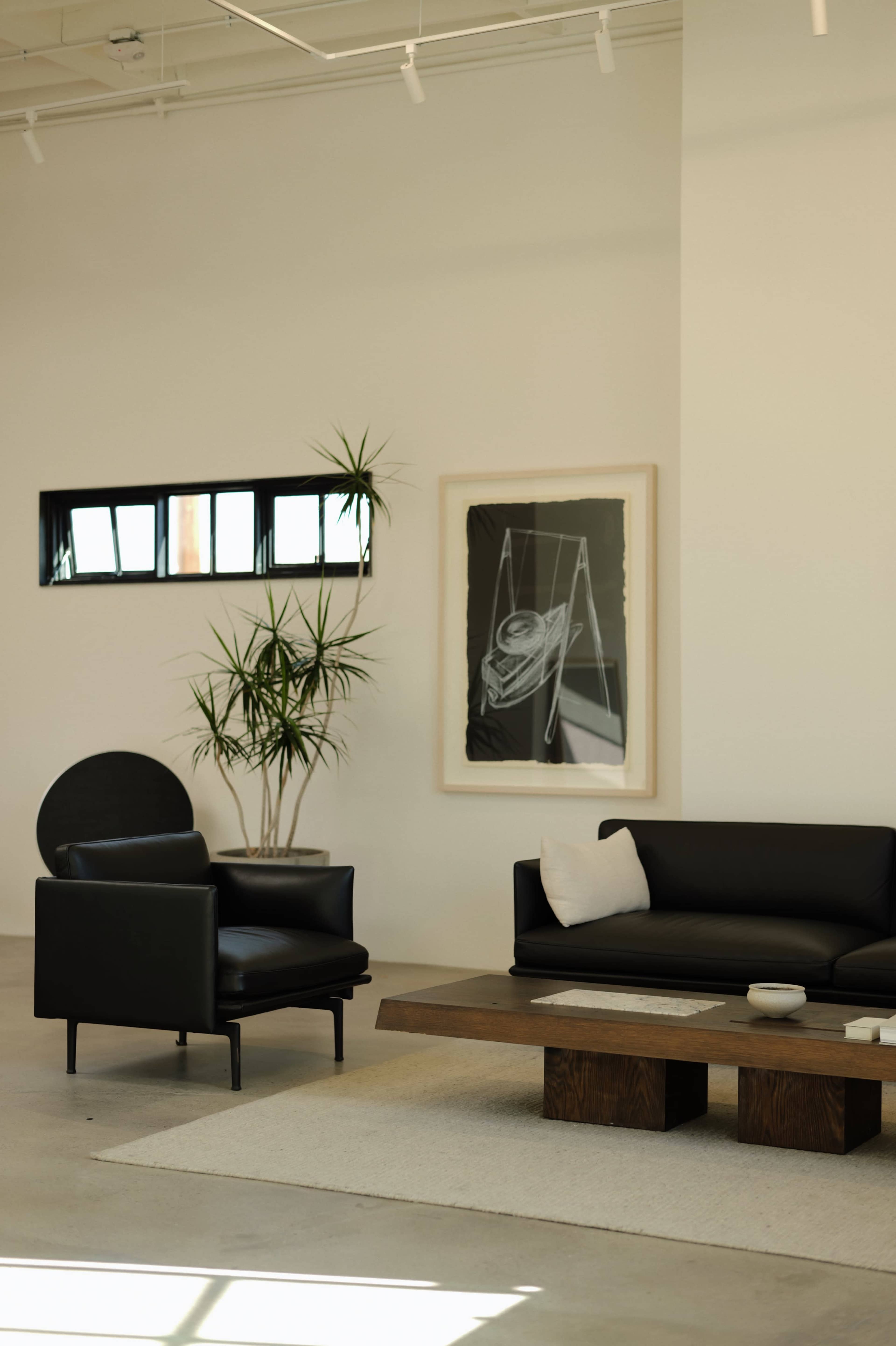 The image shows a modern living room featuring a black leather sofa and armchair, a wooden coffee table, a large framed artwork on the wall, and a plant in the corner.