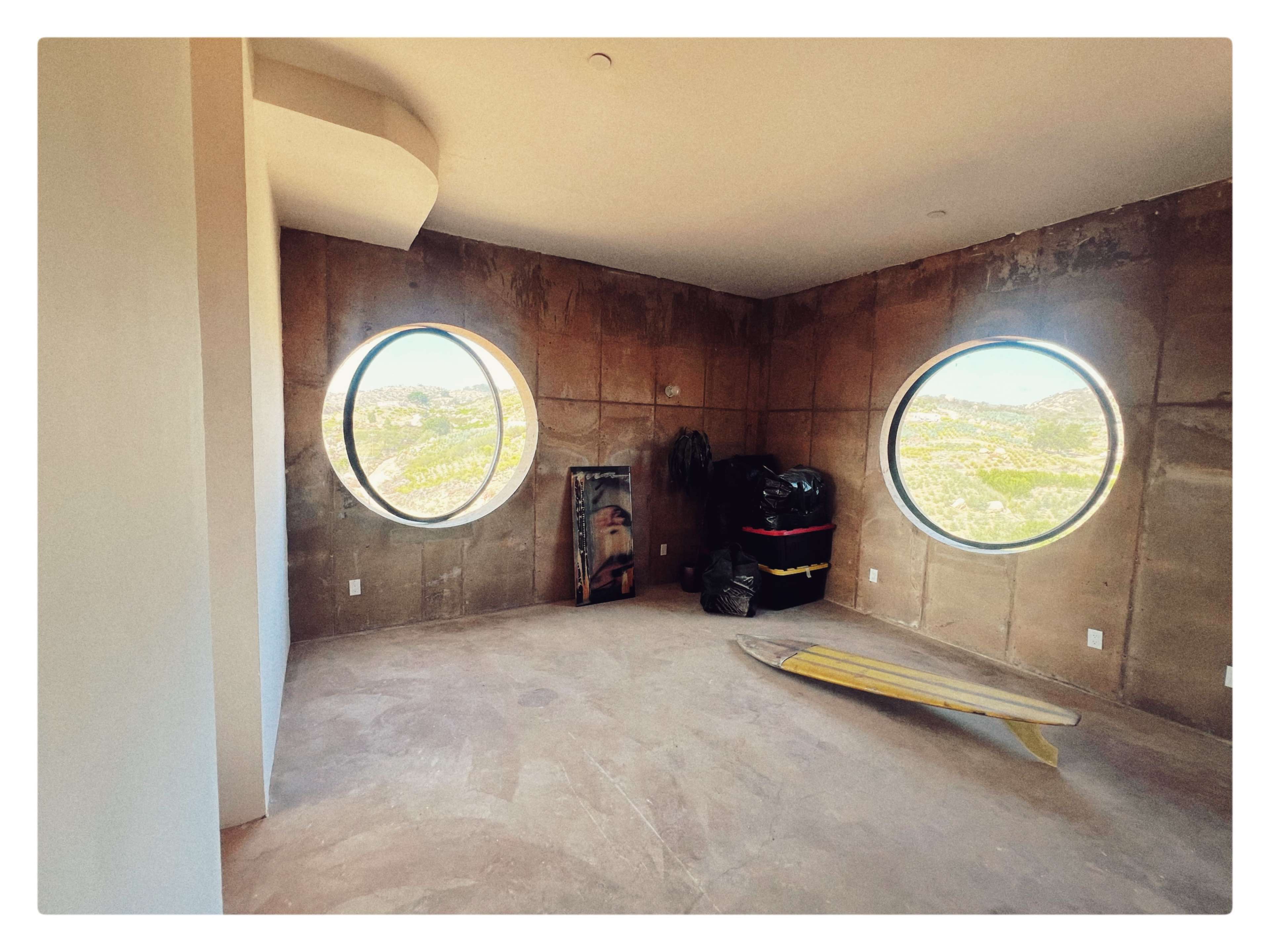 The image shows a sparsely furnished room with textured walls, featuring two large circular windows that offer a view of the outdoors.