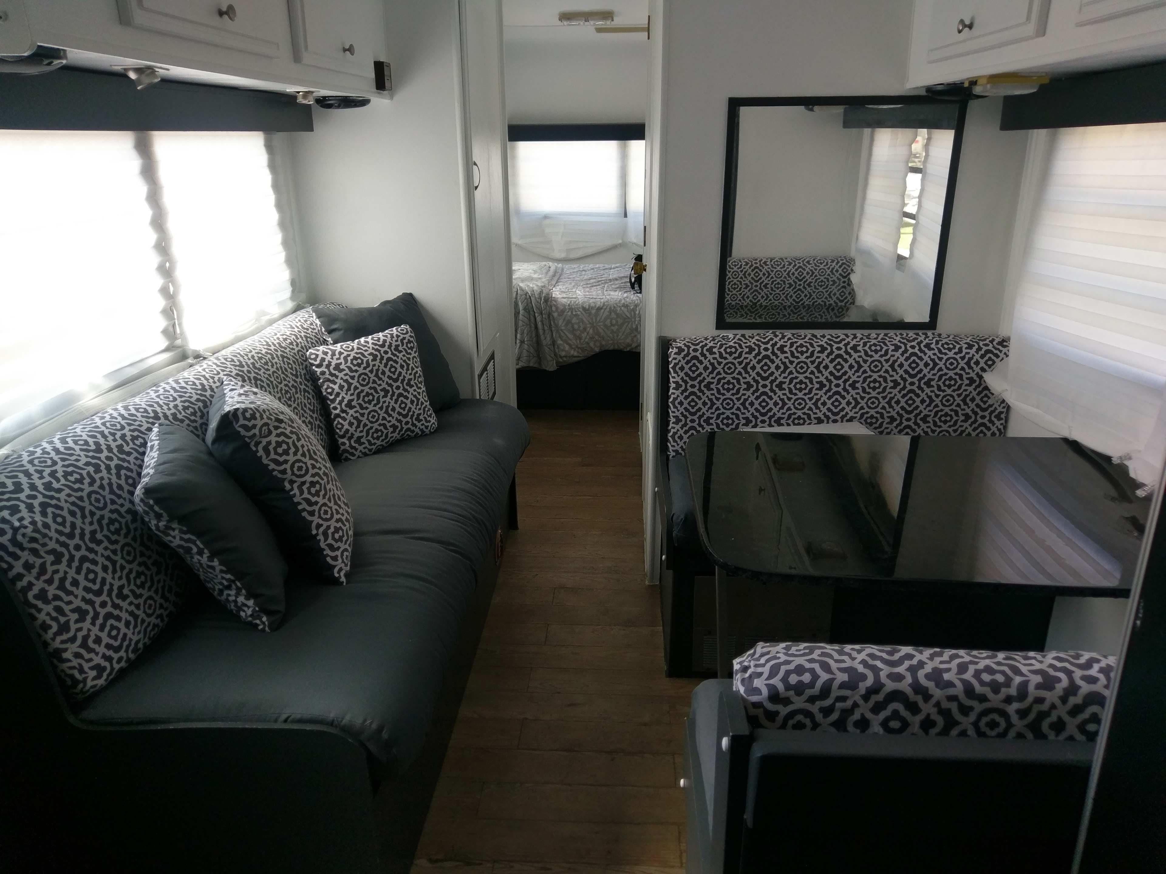 The interior of a camper trailer featuring a gray couch, patterned cushions, a black table, and a mirrored wall, with a bed visible in the rear.