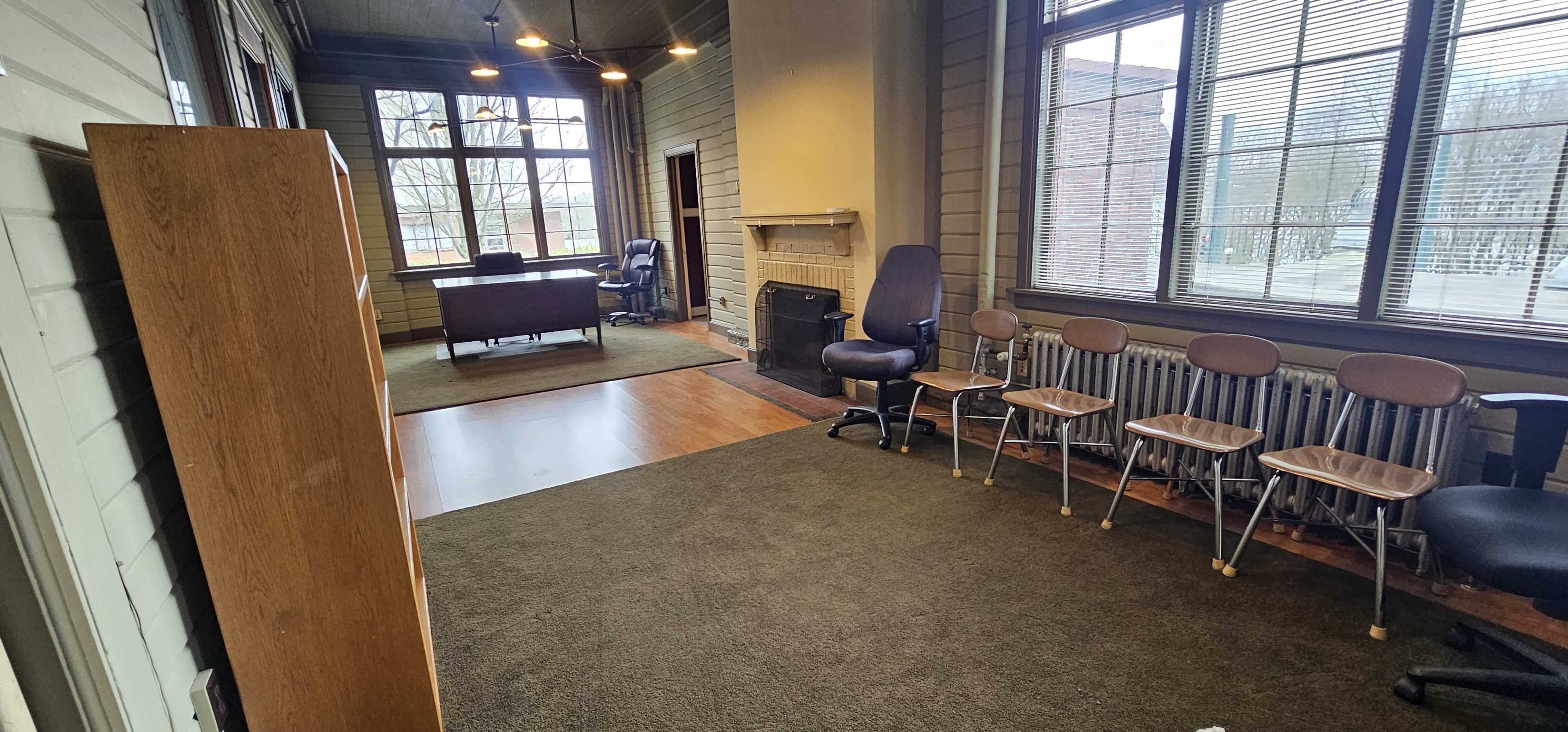A room with a desk, chairs, large windows, and a fireplace, featuring hardwood and carpeted flooring.