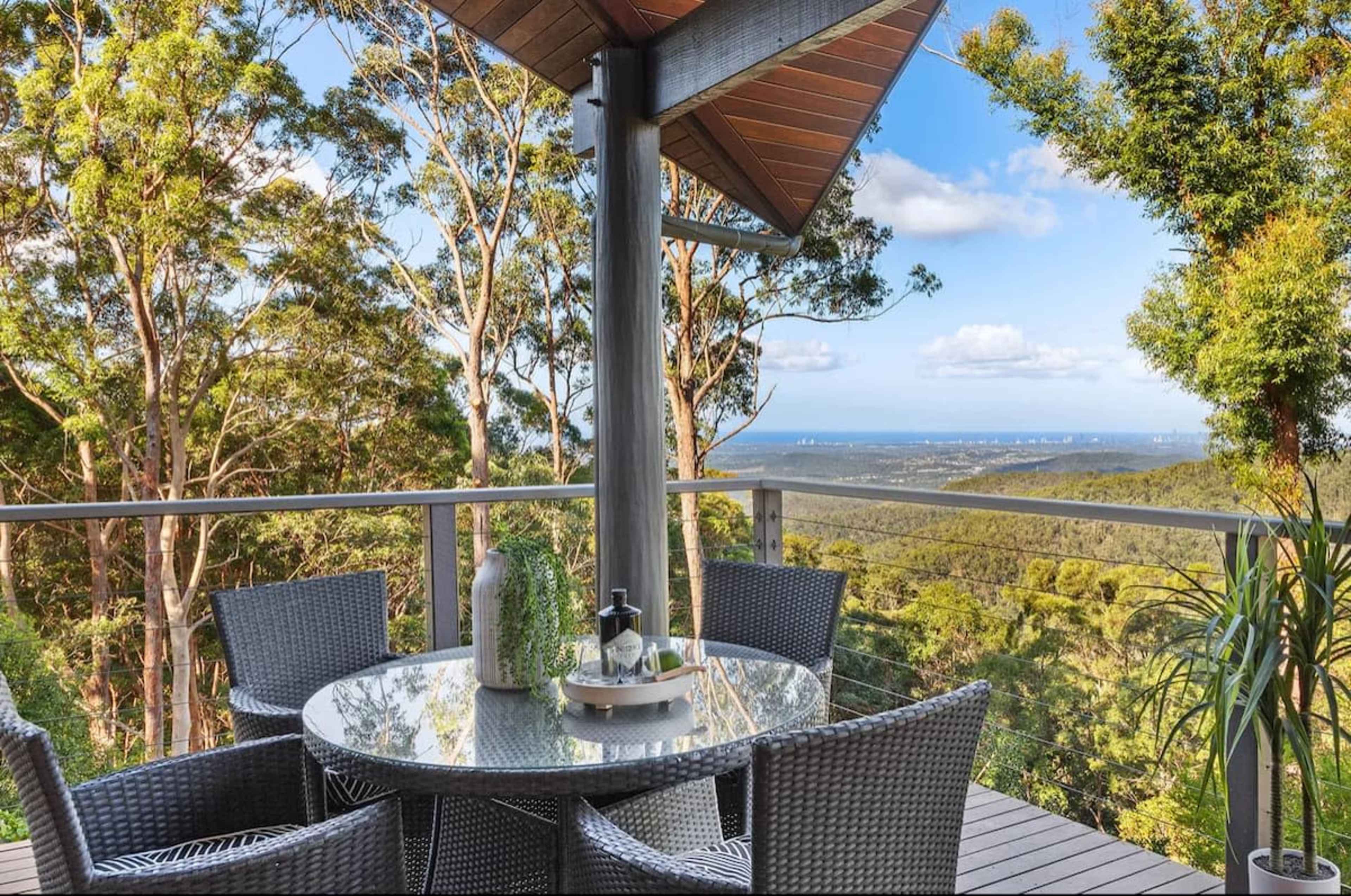 A round glass table surrounded by four chairs is set on a balcony overlooking a forested landscape and distant coastline.