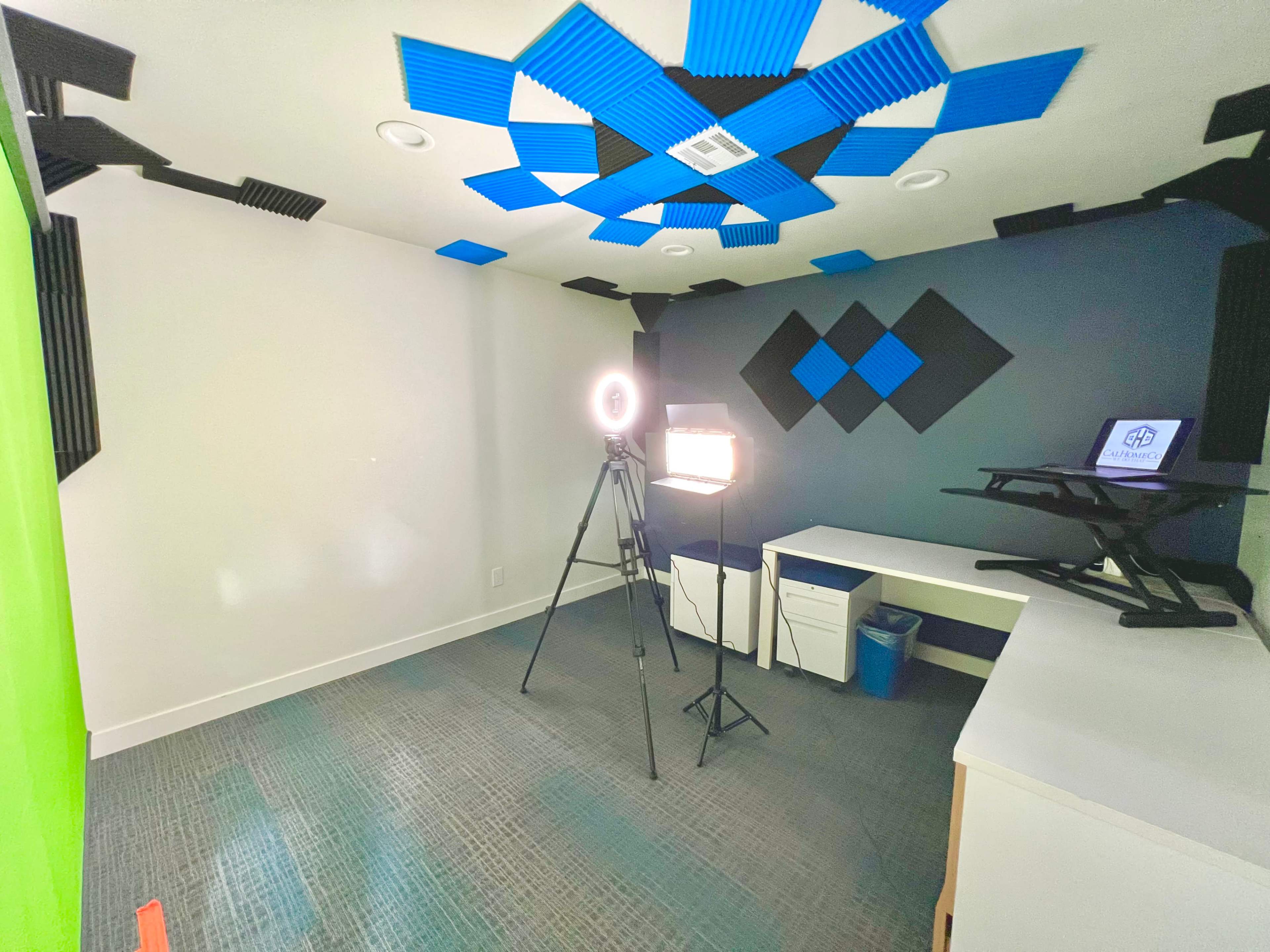 A video recording studio with a green screen, lighting setup, and a camera on a tripod facing a desk with a laptop.