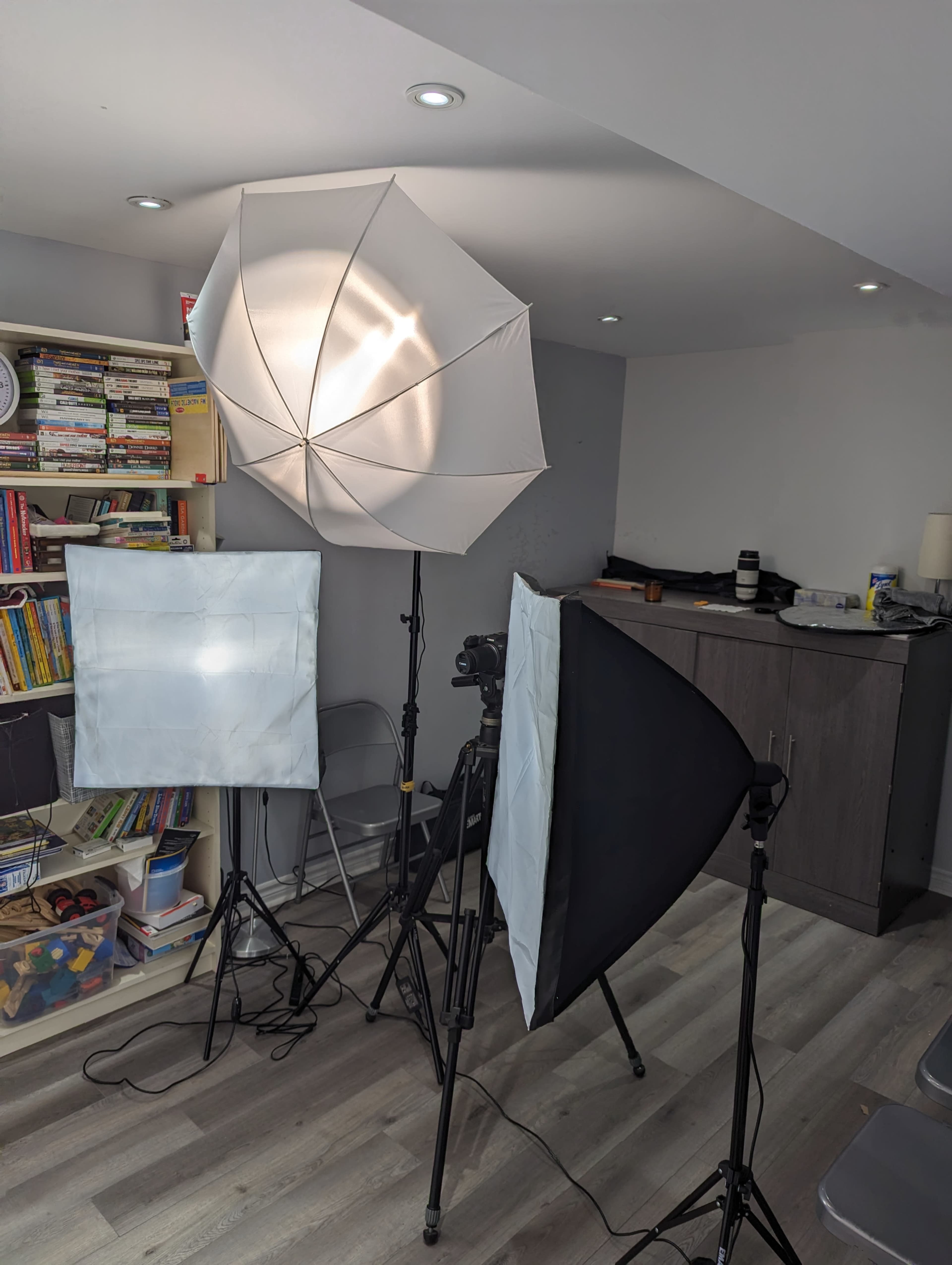 A photography setup with two softbox lights and a black backdrop positioned in a room with shelves and a gray wall.