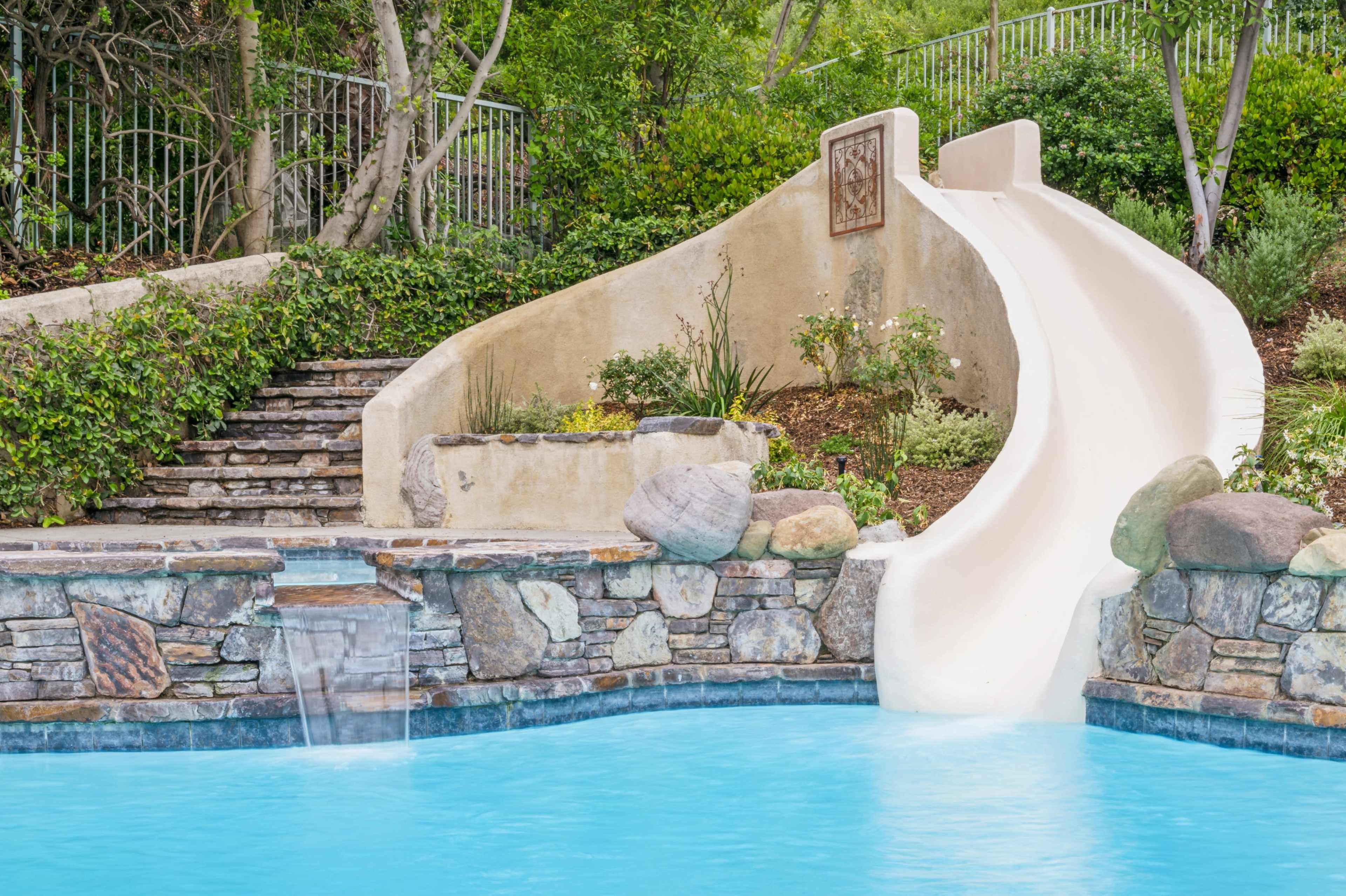 A white waterslide curves down into a blue swimming pool surrounded by stonework and greenery.
