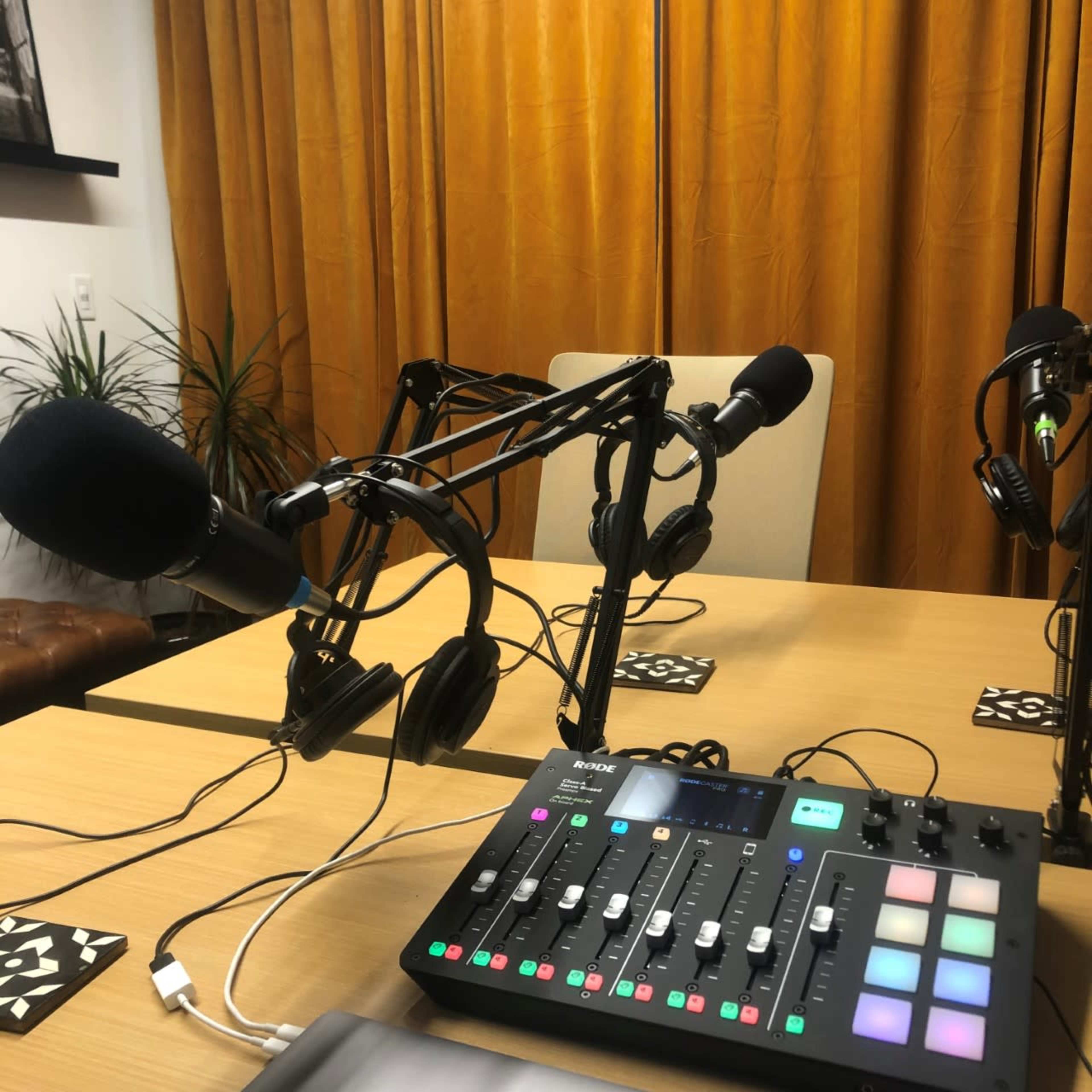 A podcasting setup with two microphones, headphones, and a mixing console on a table, all set against a backdrop of orange curtains.