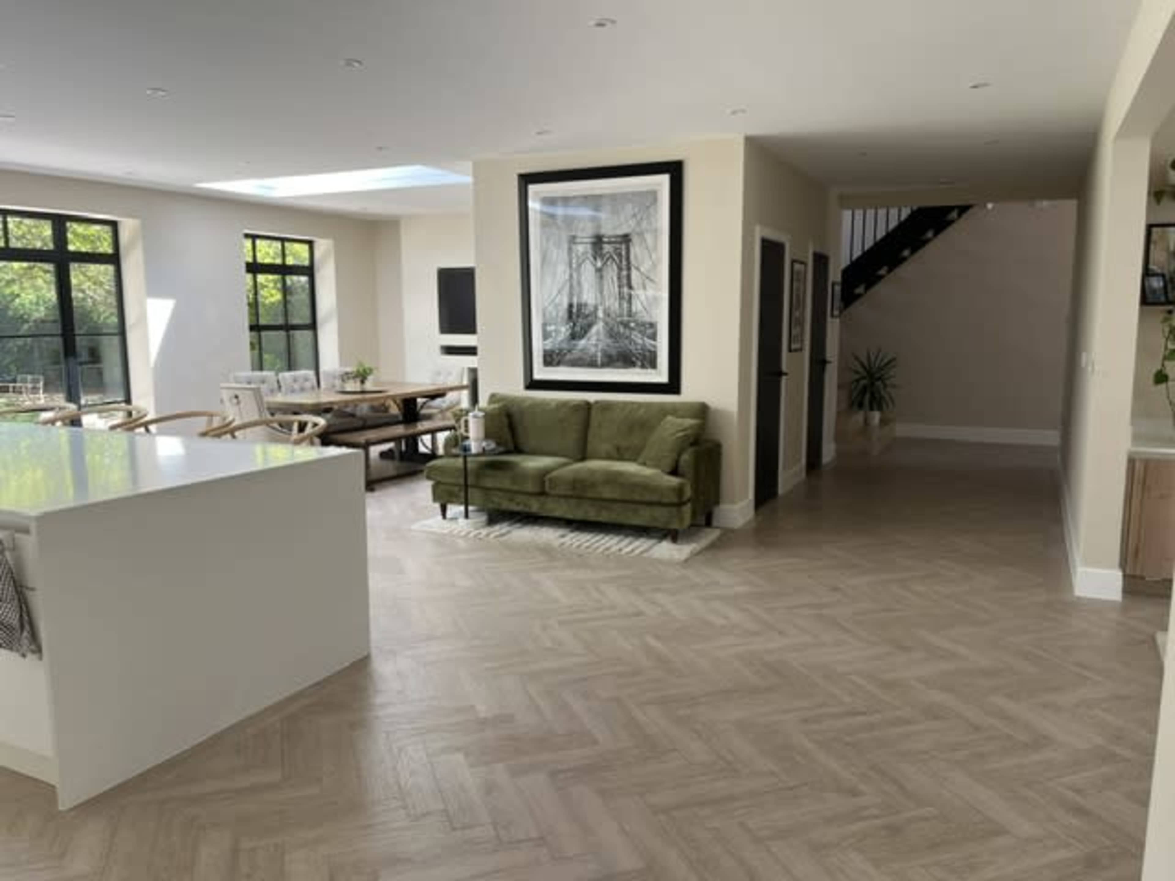 The image shows a modern open-plan living space featuring a green sofa, a dining area with a wooden table, and large windows that allow natural light to enter.