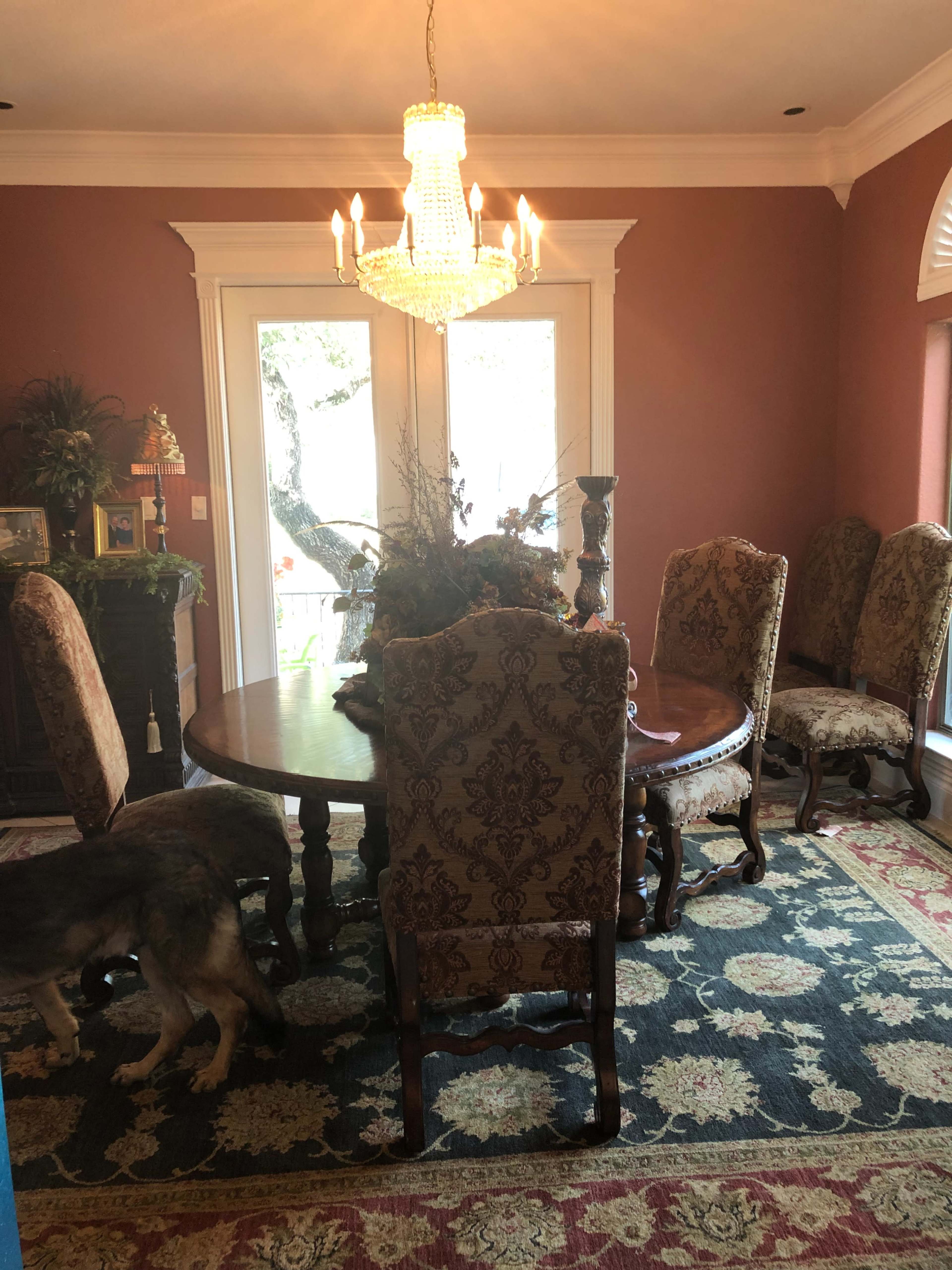 A round table surrounded by six upholstered chairs is positioned in a well-lit dining room with a chandelier and doors leading to an outdoor view.