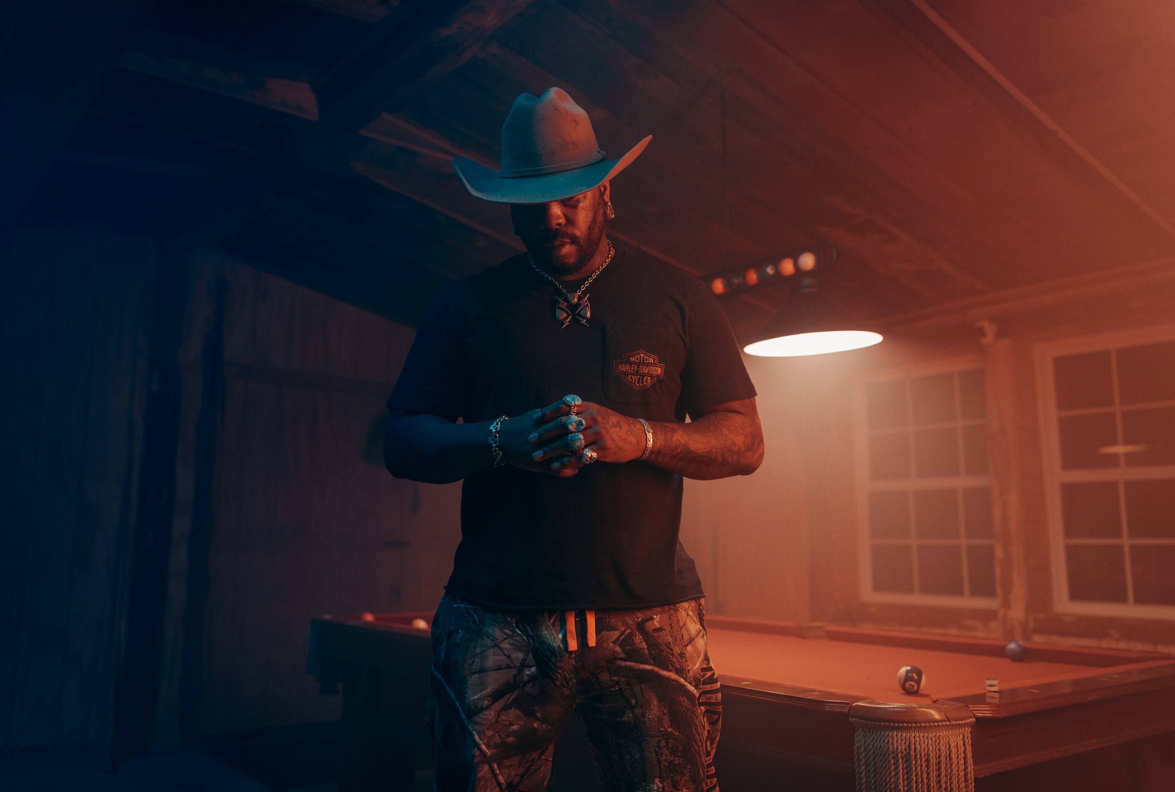 A man wearing a hat stands in a dimly lit room with a pool table.
