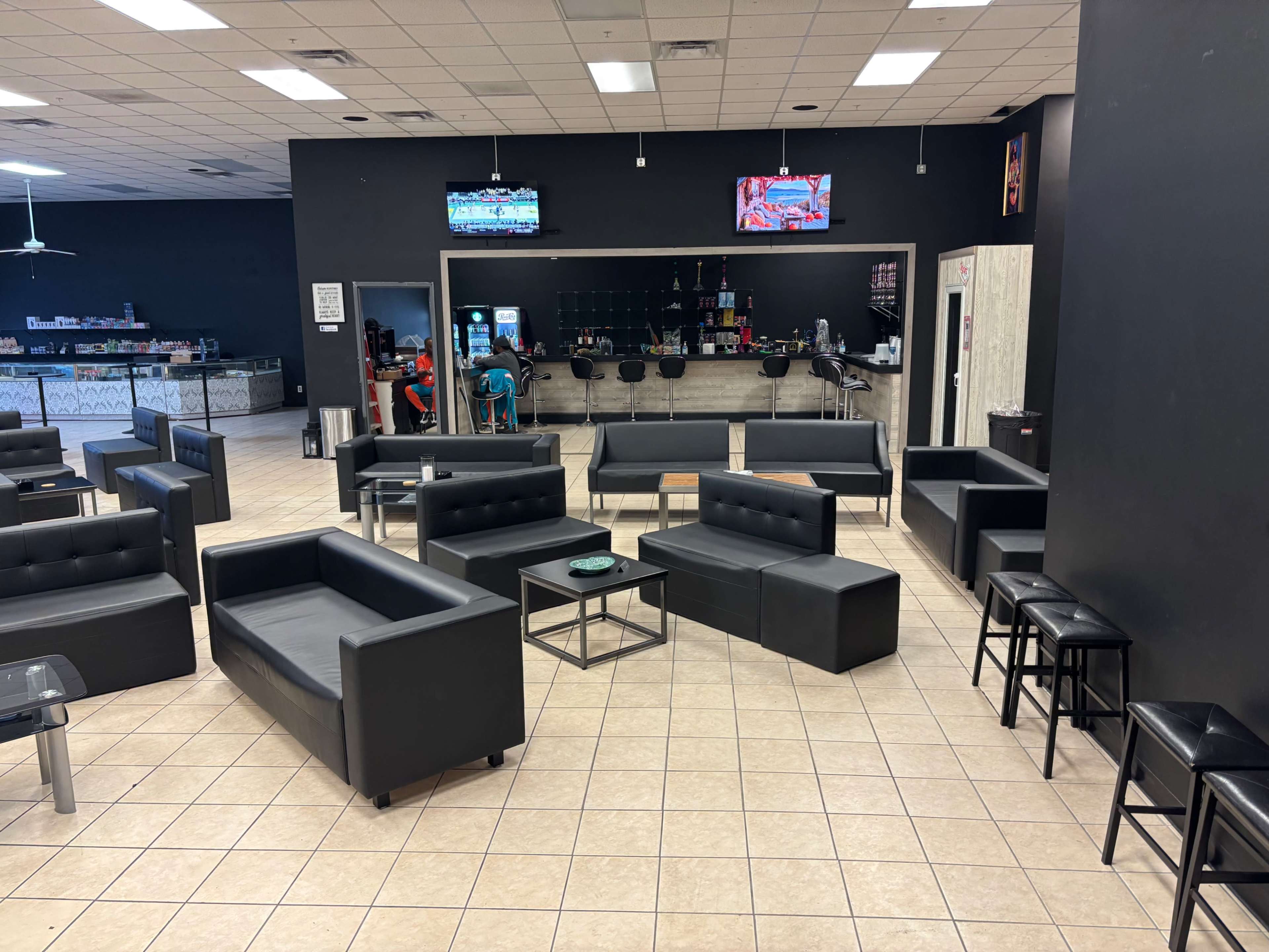 The image shows a lounge area with black couches and chairs arranged around small tables, with a bar in the background and televisions mounted on the walls.