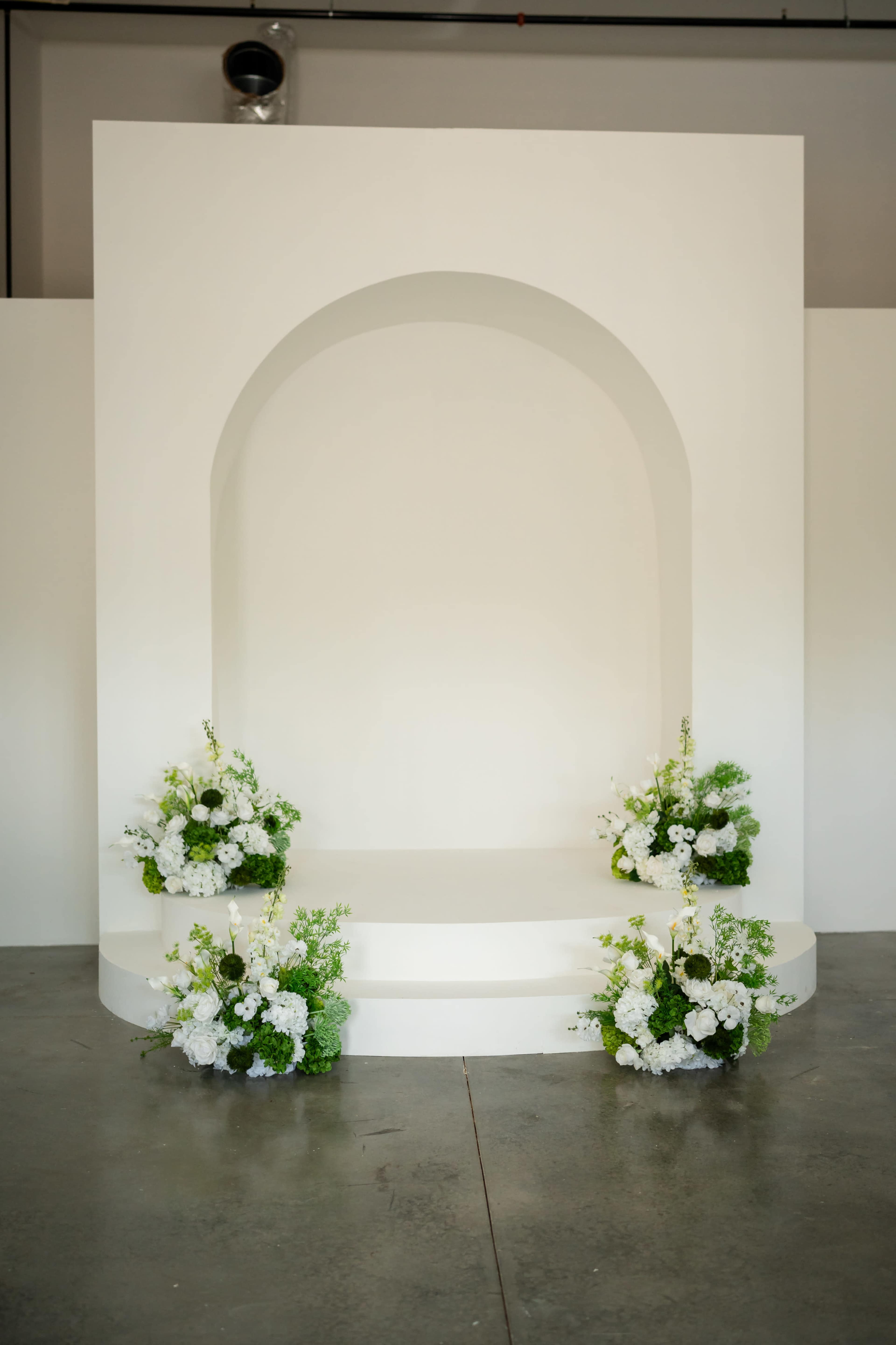 The image shows a simple, white stage with a curved arch at the center, flanked by floral arrangements on either side.