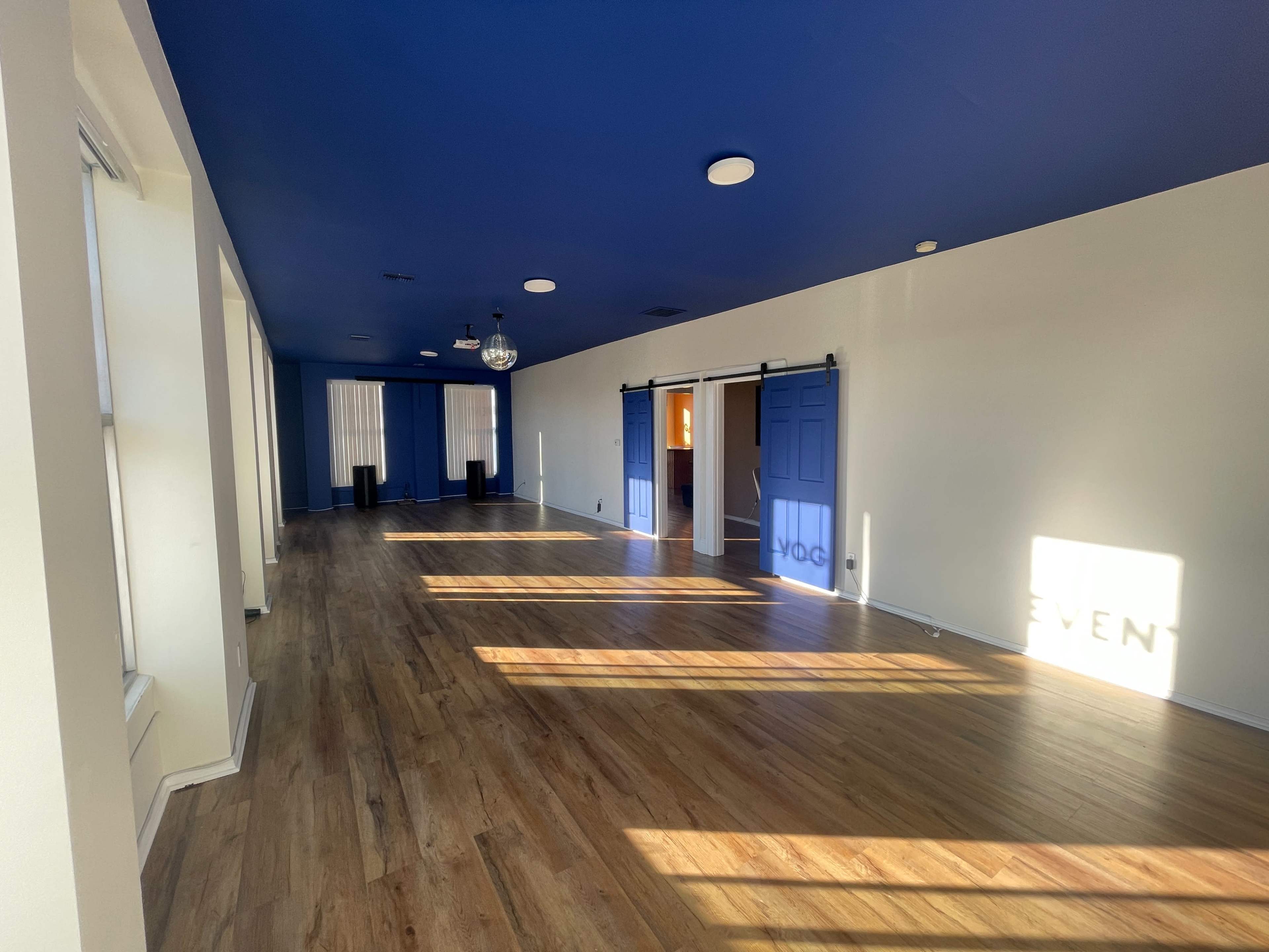 The image shows an empty room with blue painted ceilings and walls, wooden flooring, and large windows allowing natural light to enter.