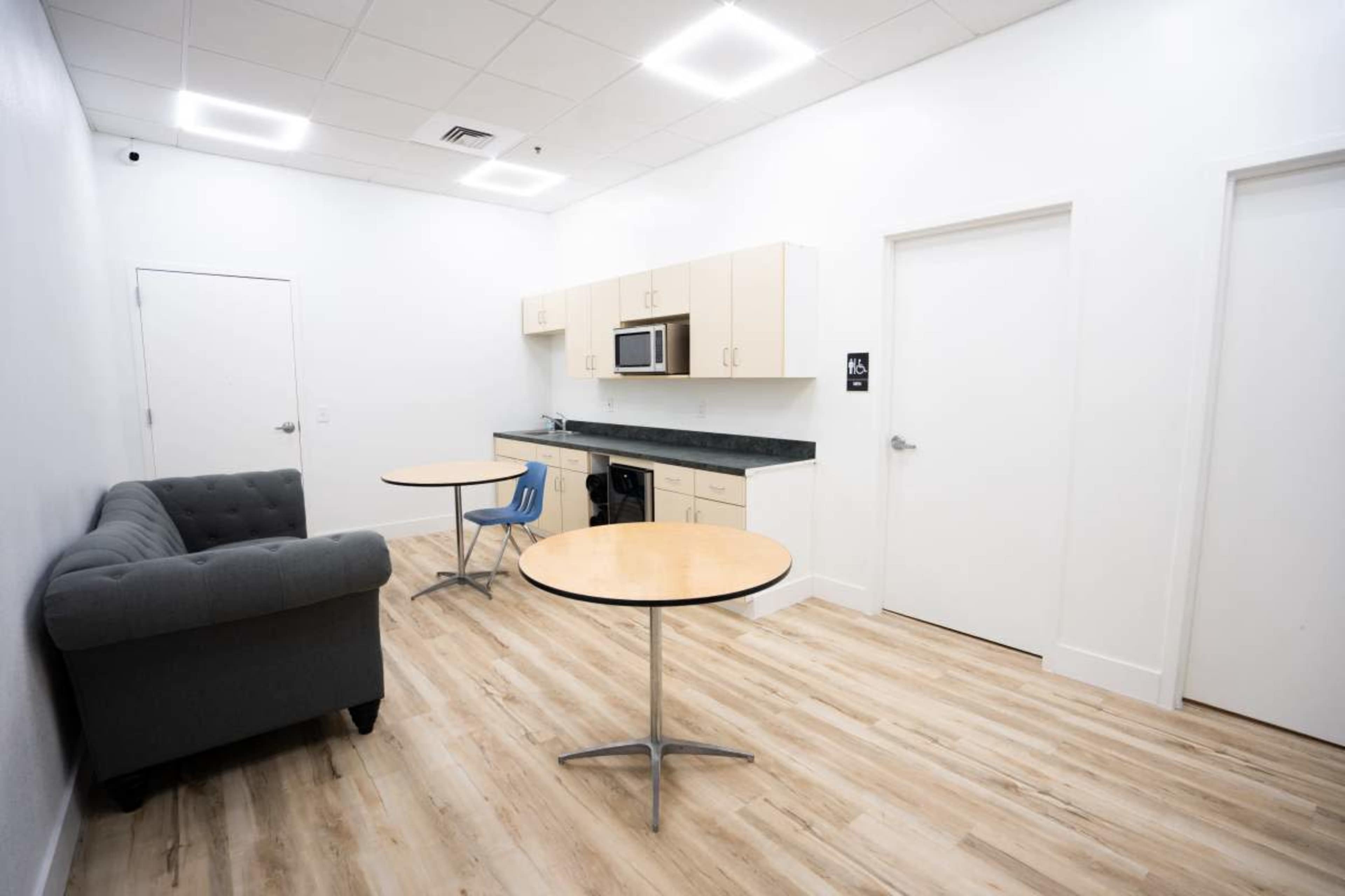 The image shows a small break room with a grey couch, two round tables, a kitchenette featuring cabinets, a microwave, and a countertop.