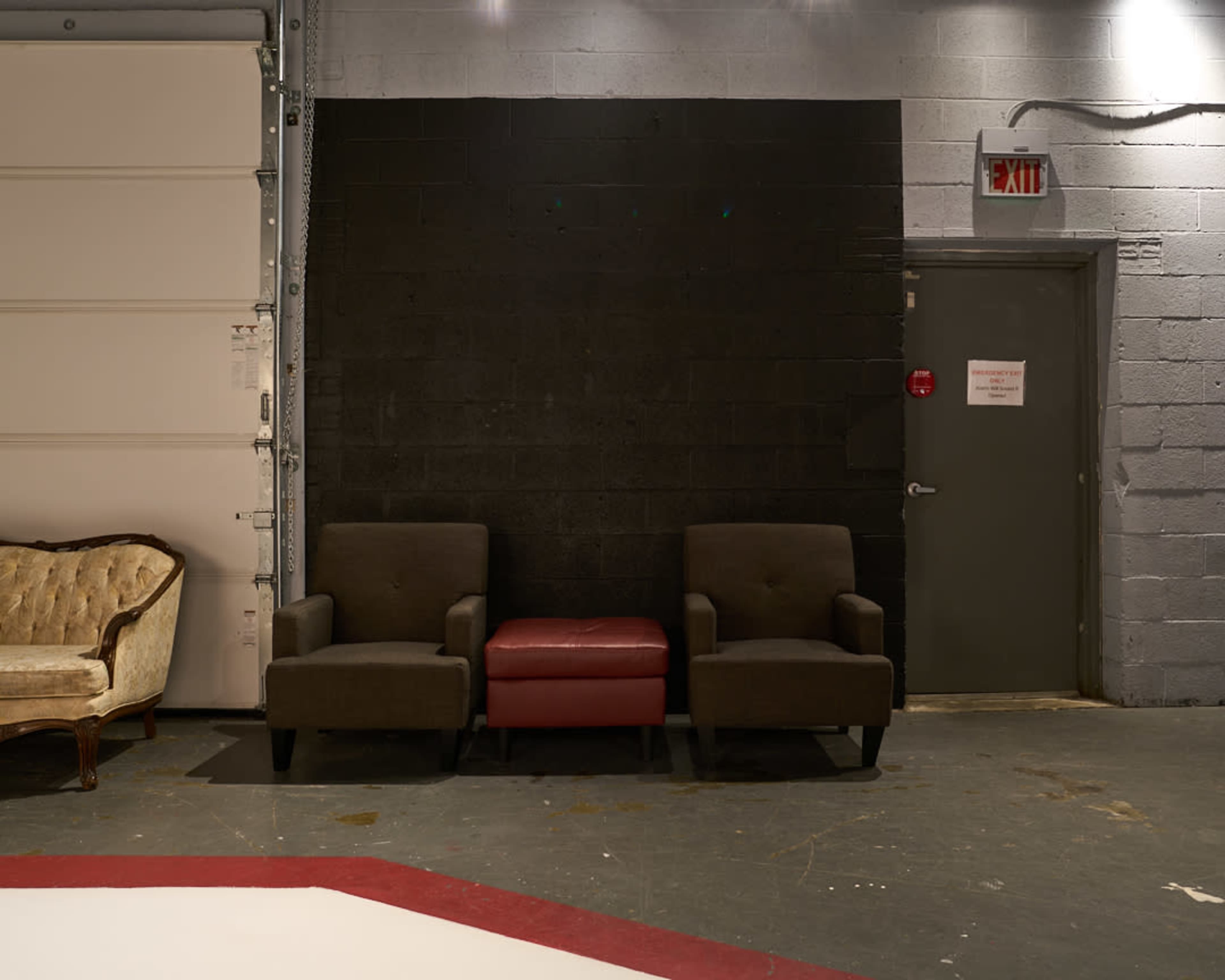 Two dark armchairs and an ottoman are positioned against a black wall in a room with a concrete floor.