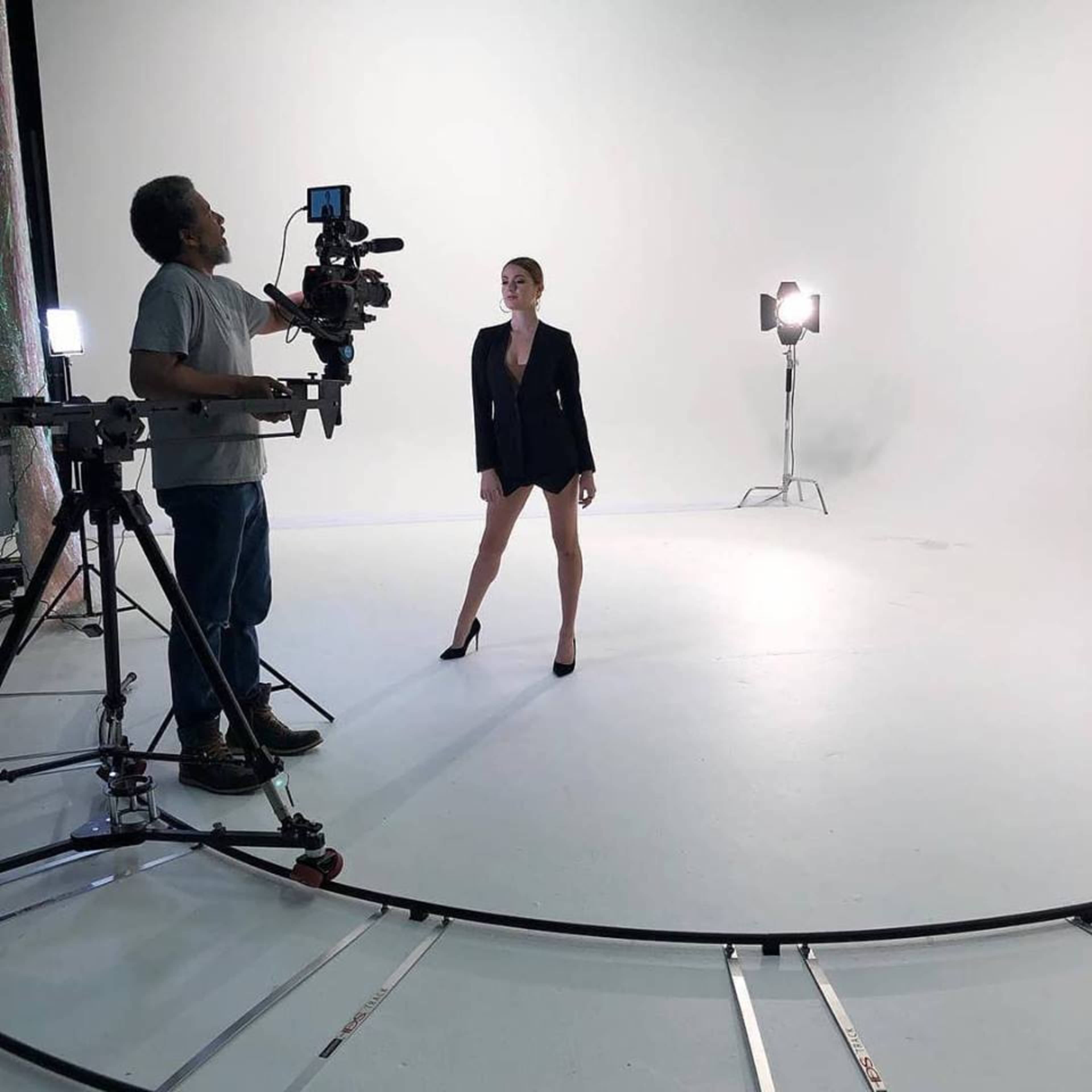 A person in a black blazer and high heels poses in a well-lit studio while a camera operator adjusts equipment.