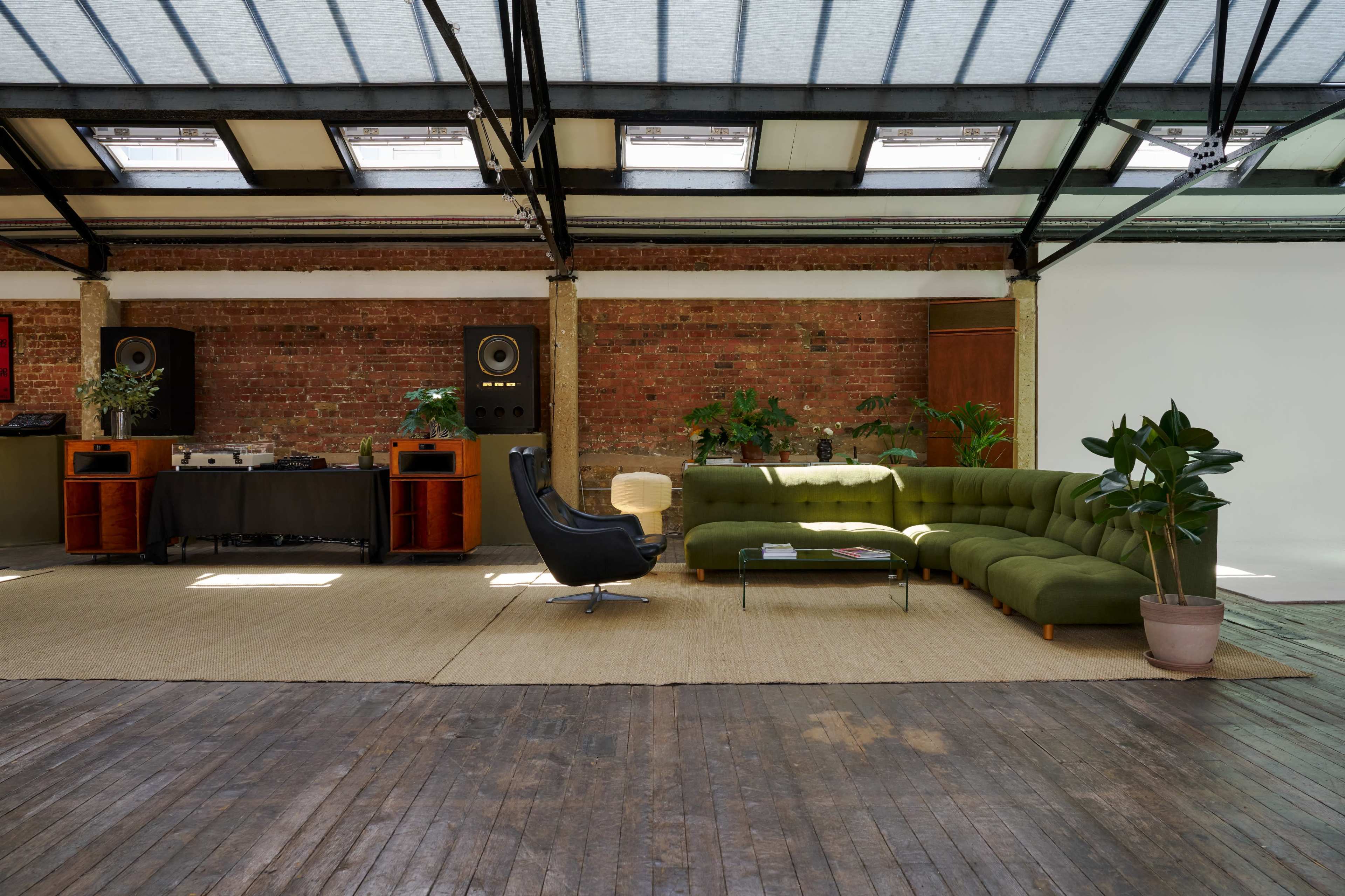 The image shows a spacious, modern living area with a green sectional sofa, a black lounge chair, and wooden floors, illuminated by natural light from large skylights.