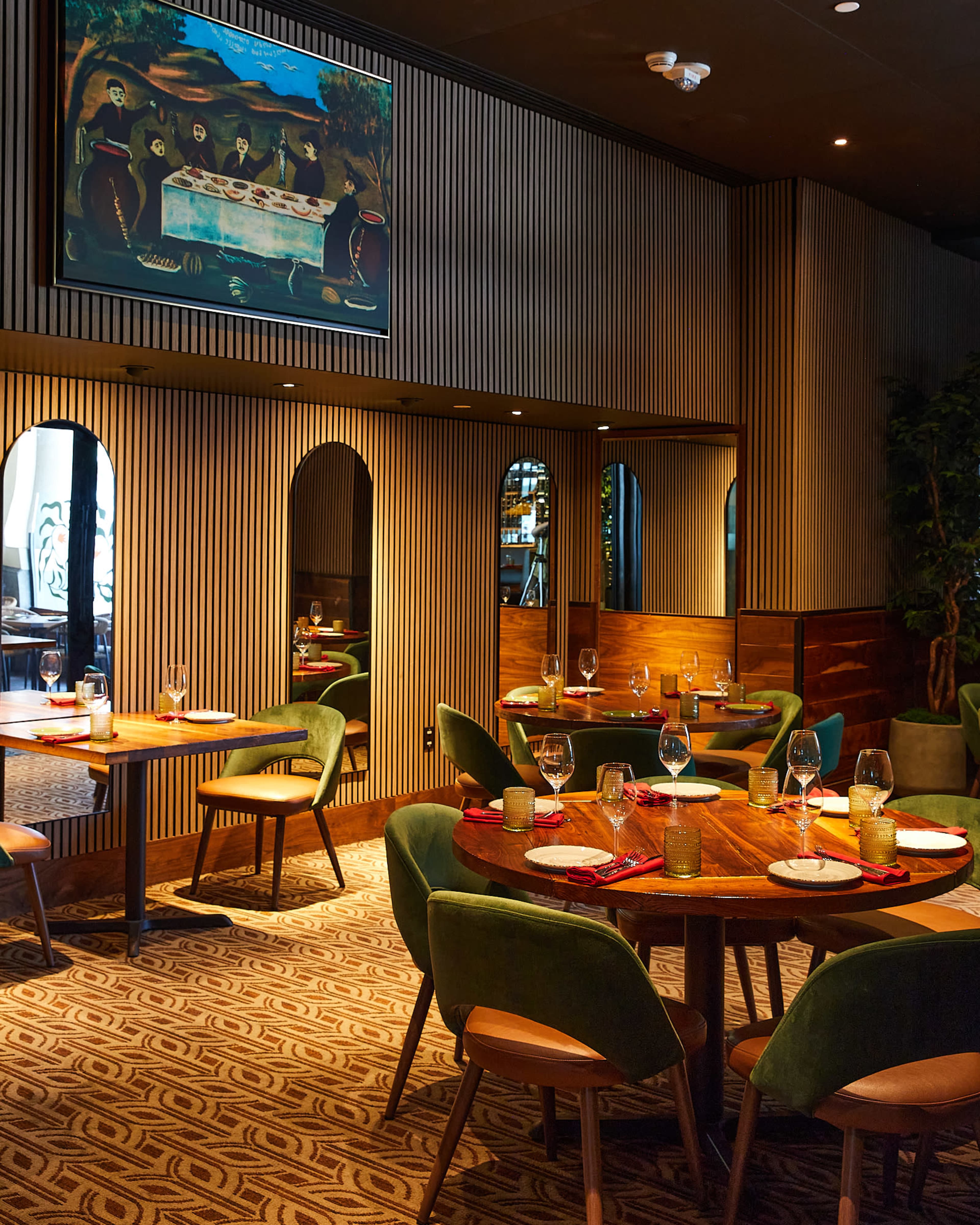 The image features a stylish restaurant interior with round wooden tables set for dining, green upholstered chairs, and a colorful painting hanging on the wall.