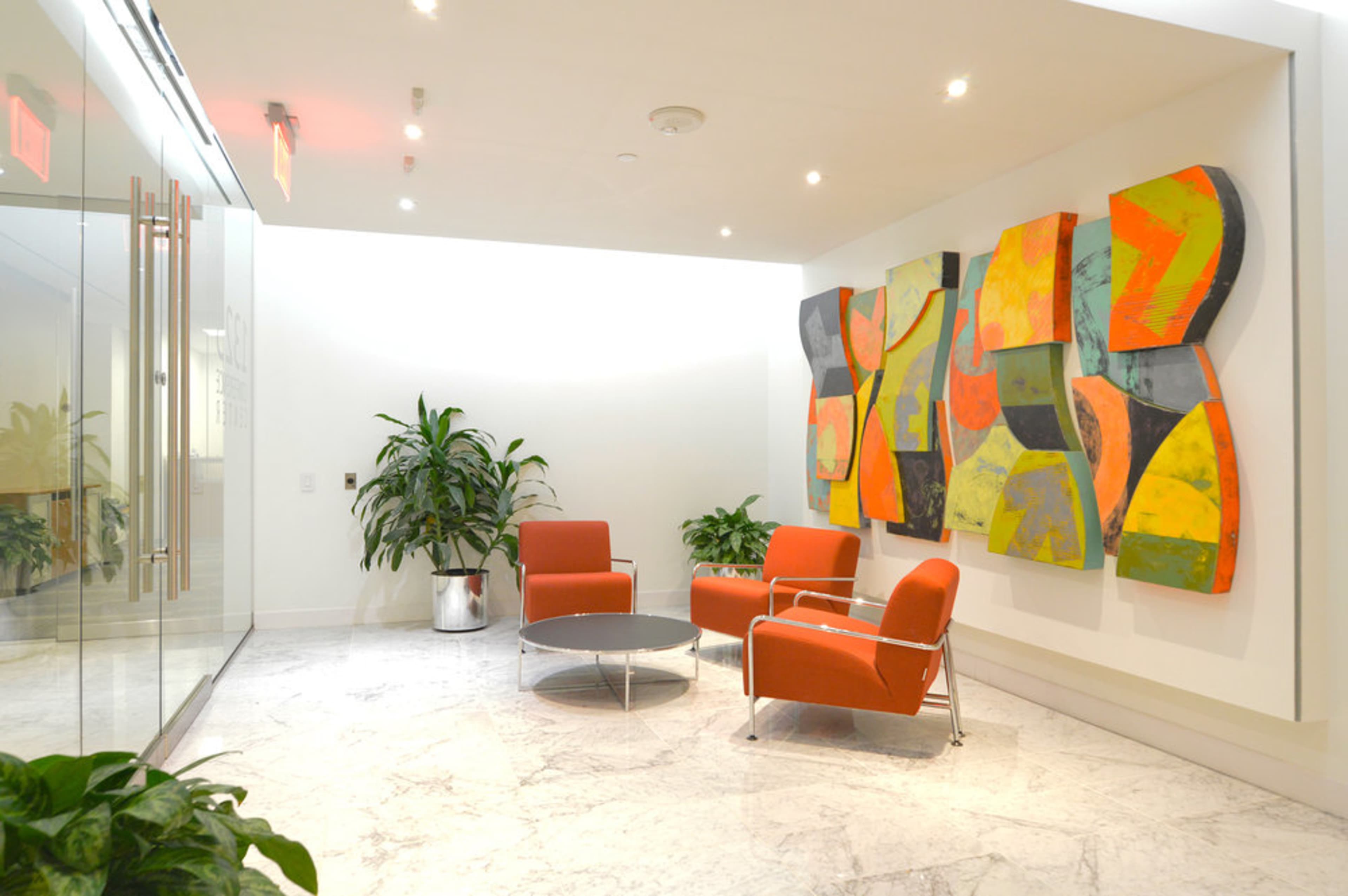 A modern waiting area with orange chairs, a circular table, and a large abstract painting on the wall.