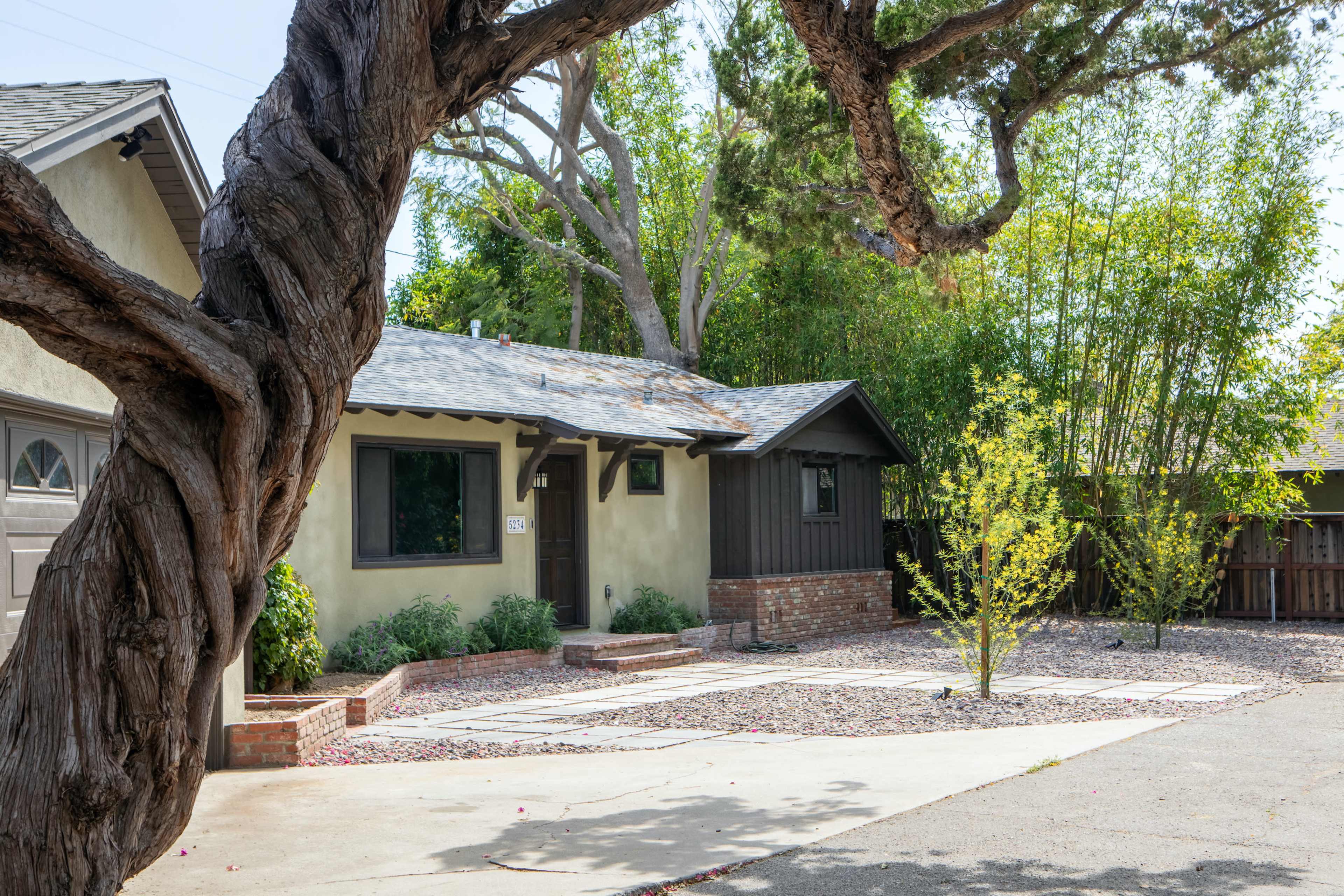 A single-story house with a gray roof, surrounded by brick landscaping and small trees, sits next to a gravel parking area.