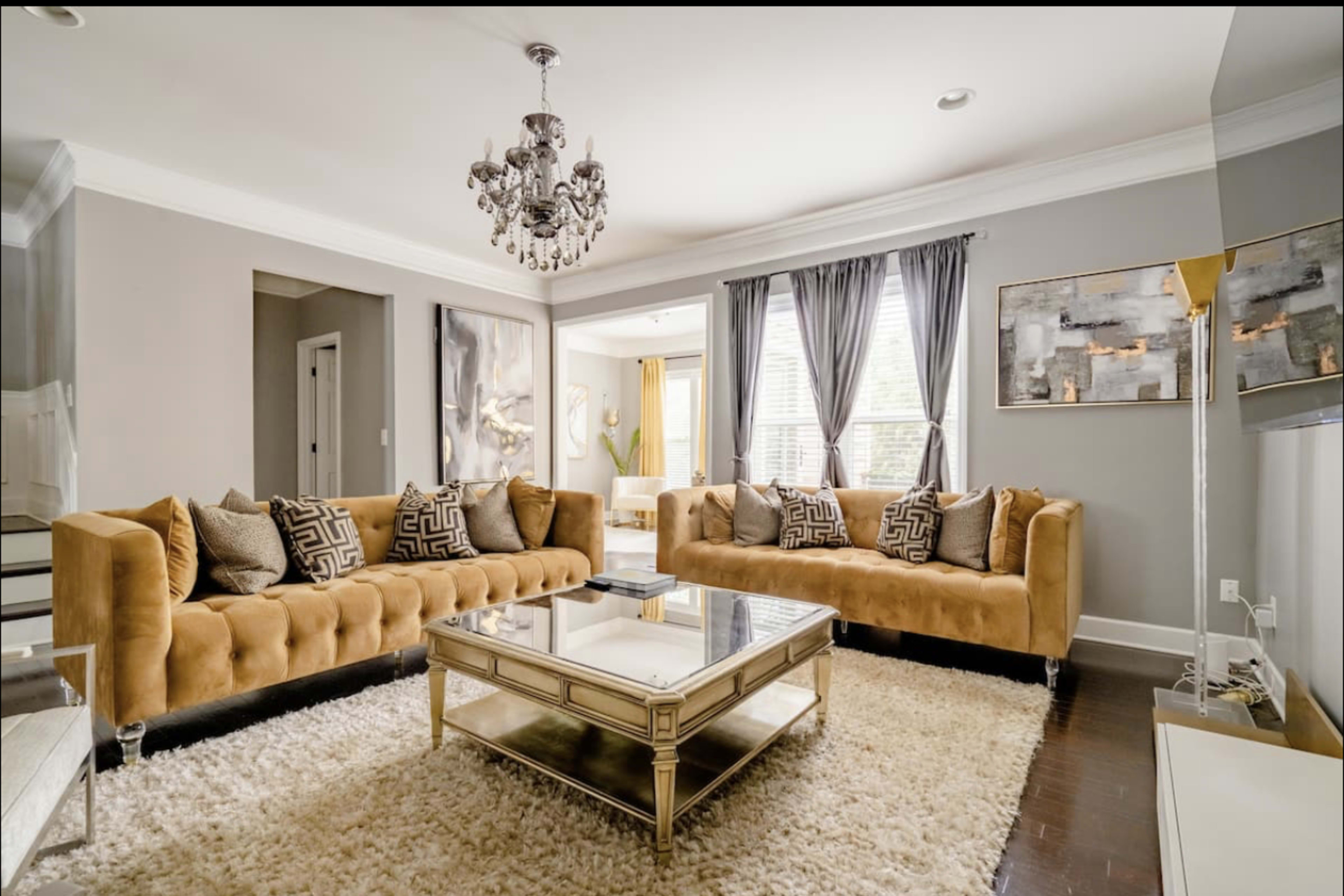 The living room features a beige tufted sofa arranged around a glass coffee table on a plush white area rug, with artwork and curtains enhancing the decor.