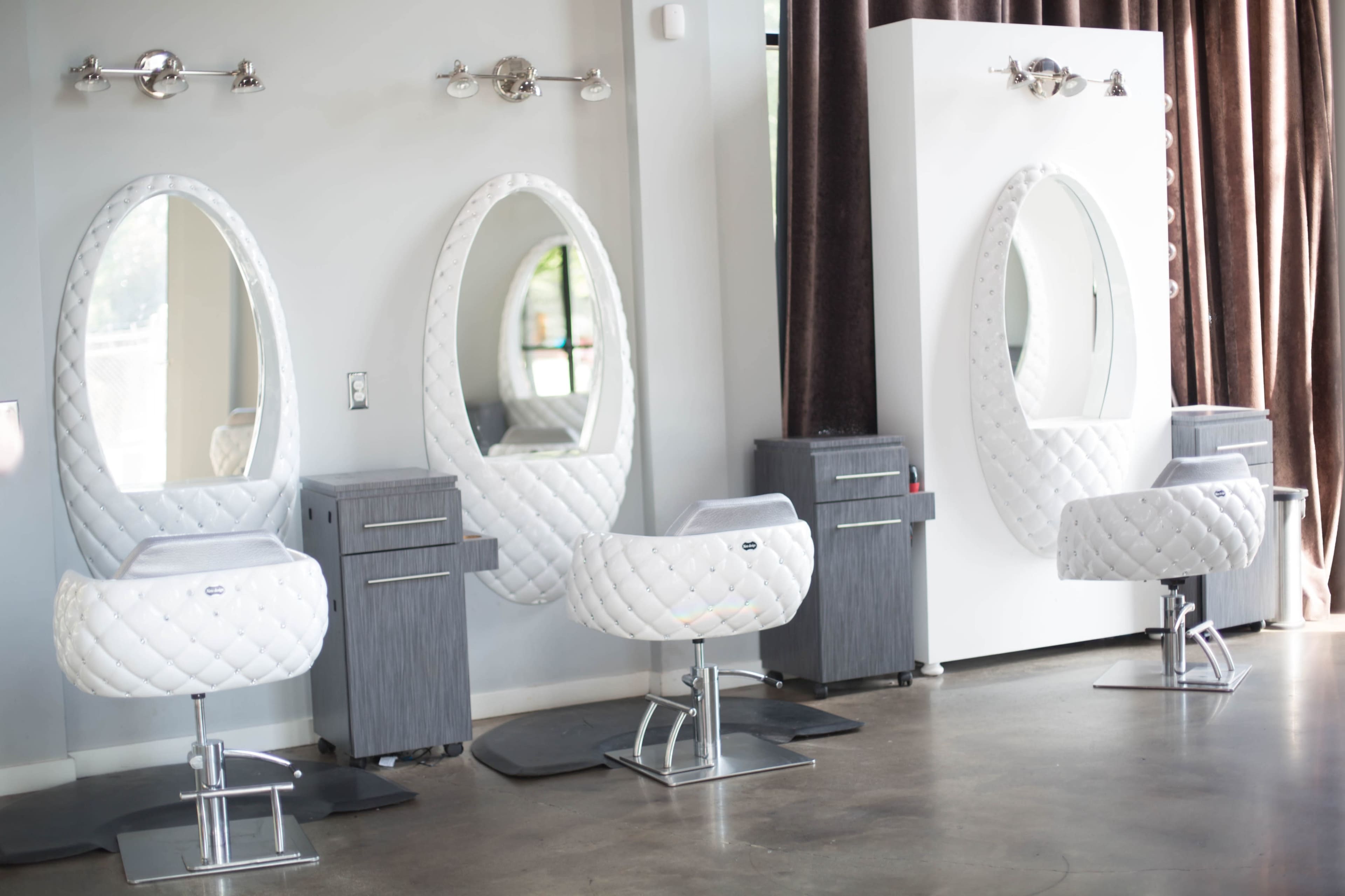 The image shows three elegantly designed salon styling stations with large oval mirrors and upholstered white chairs, set against a neutral wall and floor.