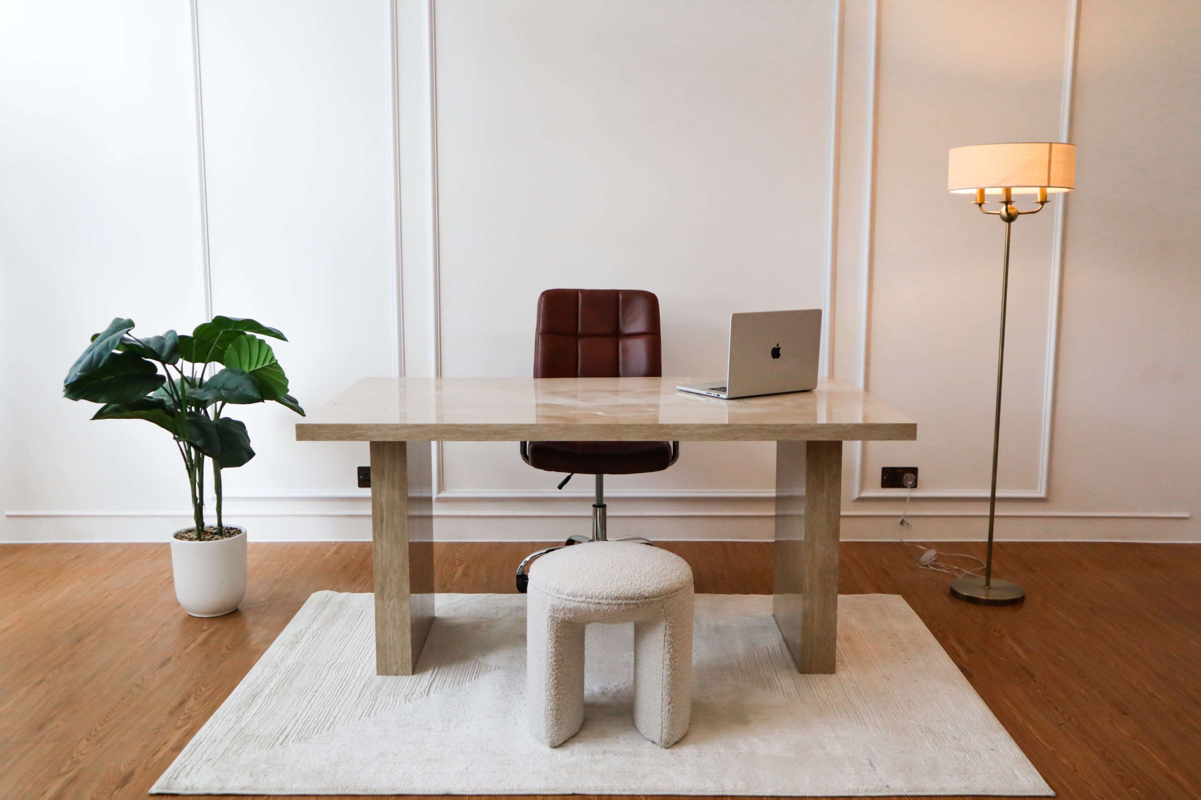 A minimalist office space features a desk with a laptop, a chair, a round cushioned stool, a floor lamp, and a potted plant.