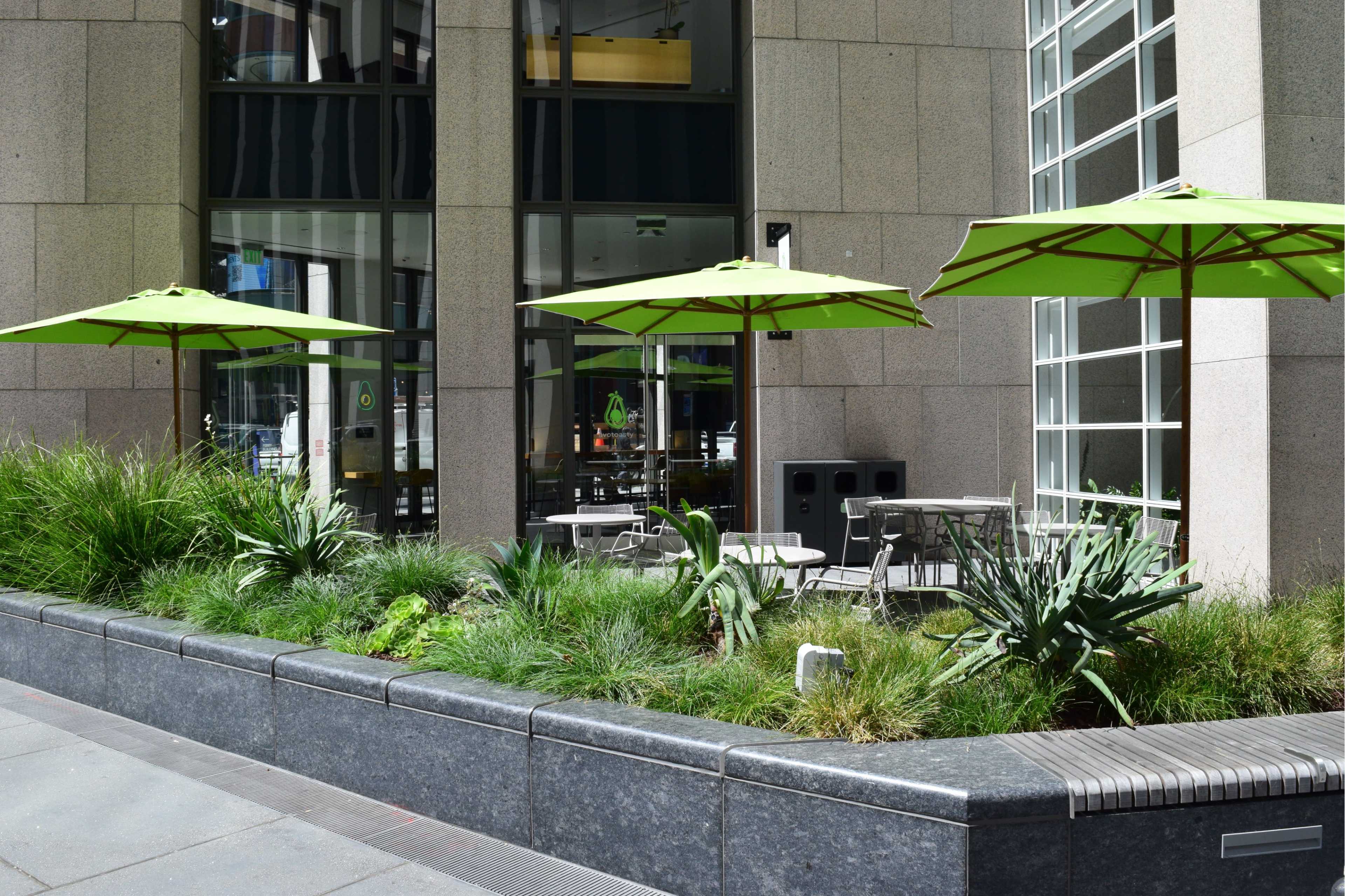 The image shows a modern outdoor seating area with green umbrellas, surrounded by flowering plants and a stone planter.