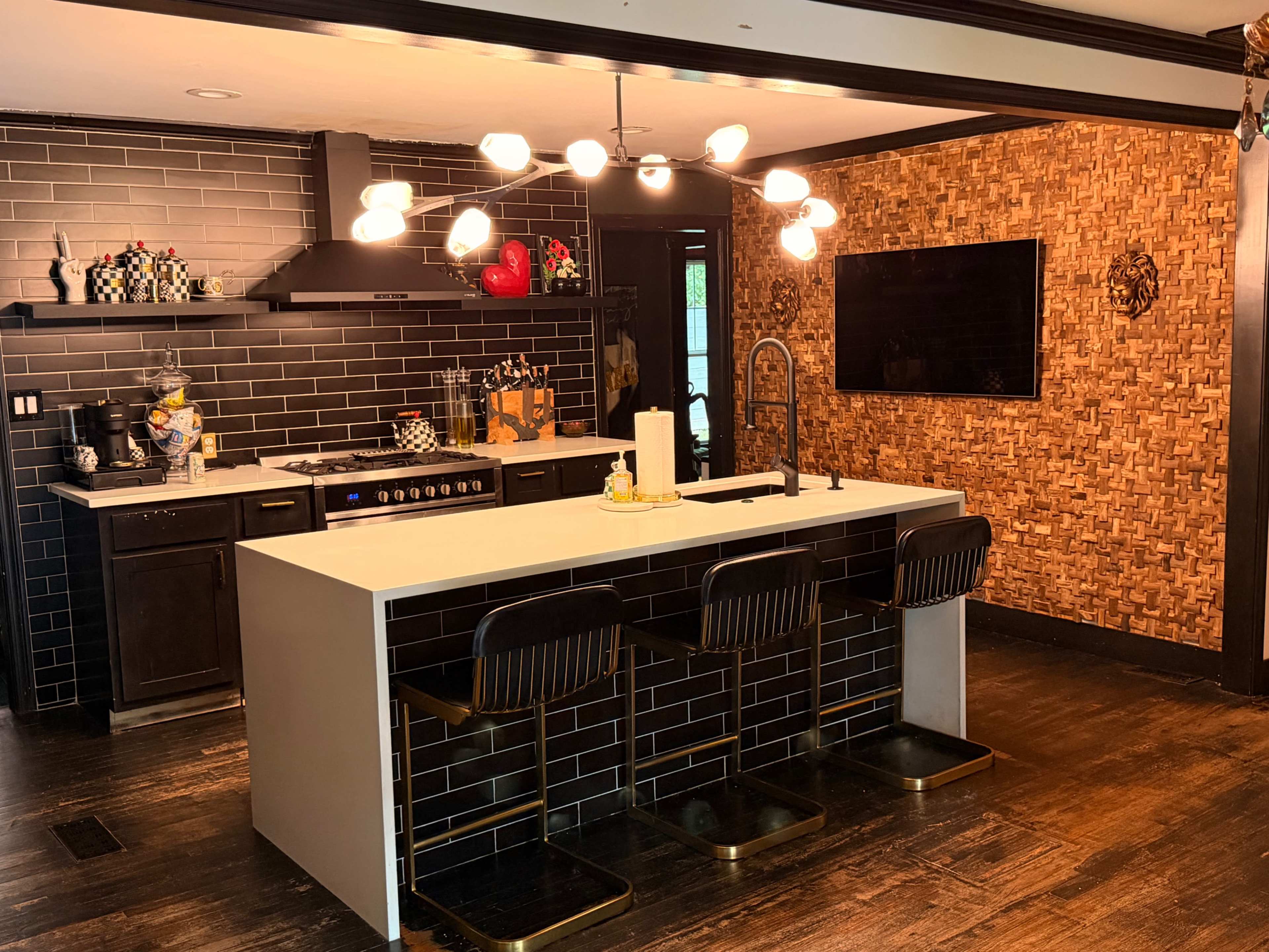 The image shows a modern kitchen featuring black tile walls, stainless steel appliances, a central island with seating, and decorative wallpaper on one side.