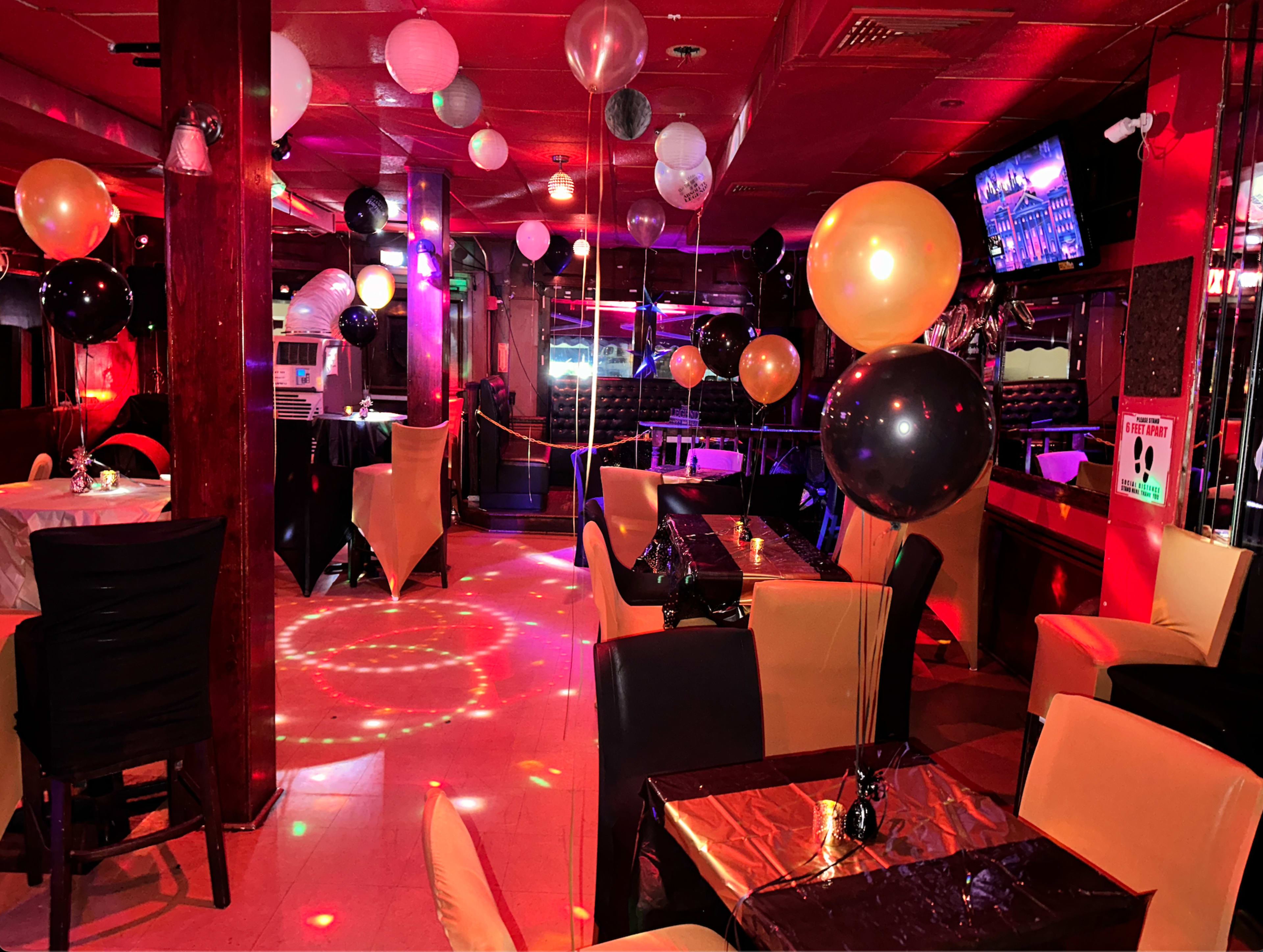 A lively restaurant interior decorated with black, white, and gold balloons, featuring tables set with dark tablecloths and colorful lighting effects on the floor.