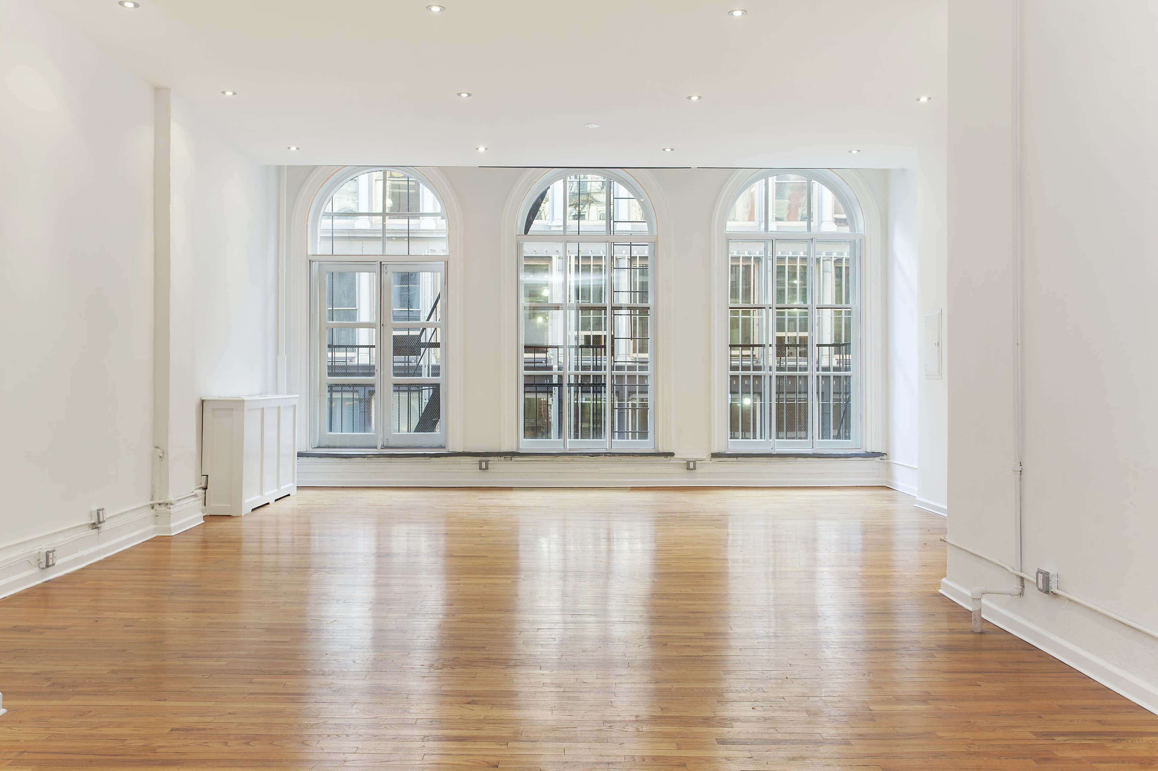 A spacious, empty room with large arched windows allowing natural light to fill the space, showcasing polished wooden floors and white walls.