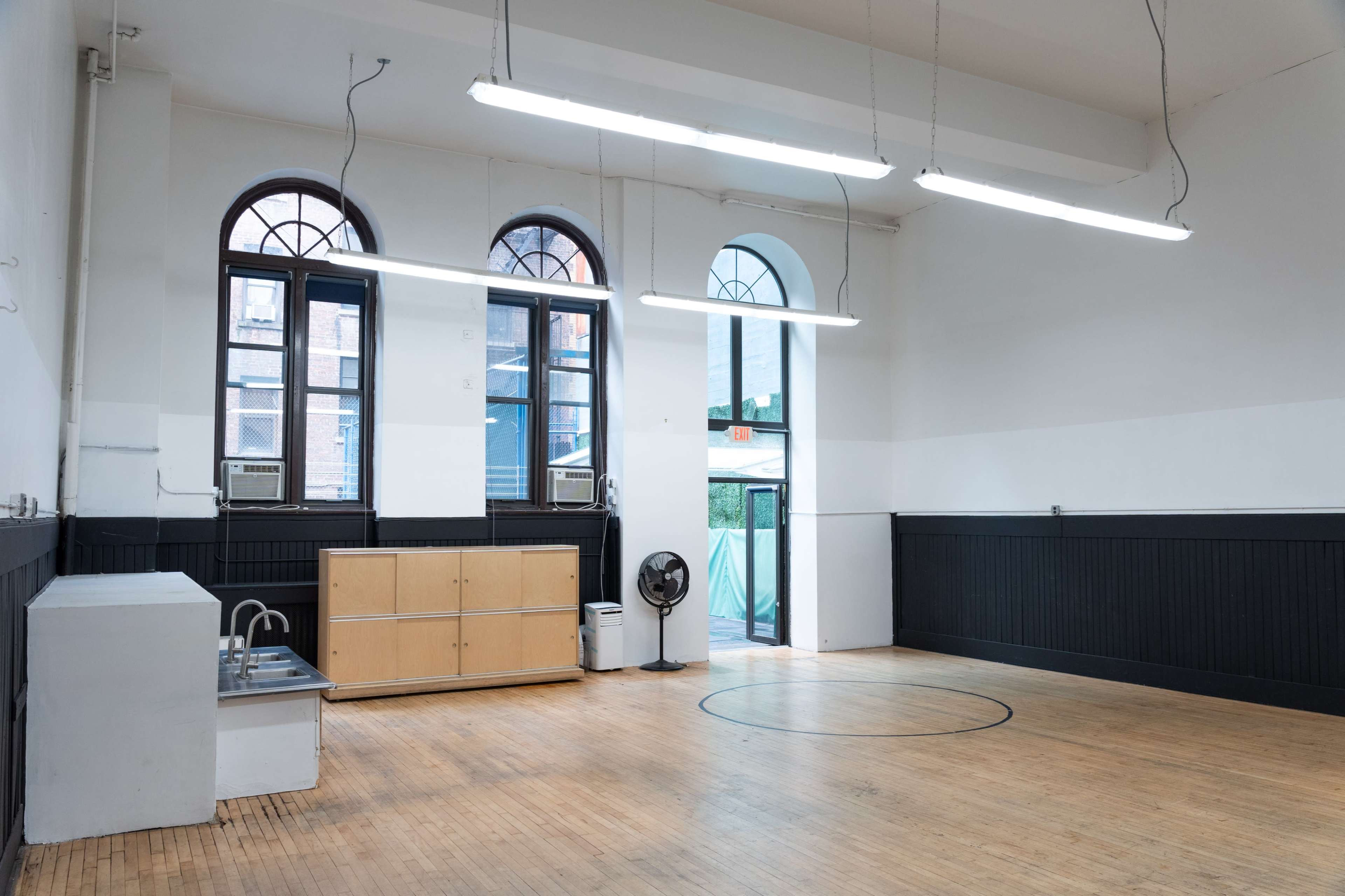 The image shows a spacious, empty room with a wooden floor, large windows, and a kitchenette against one wall.