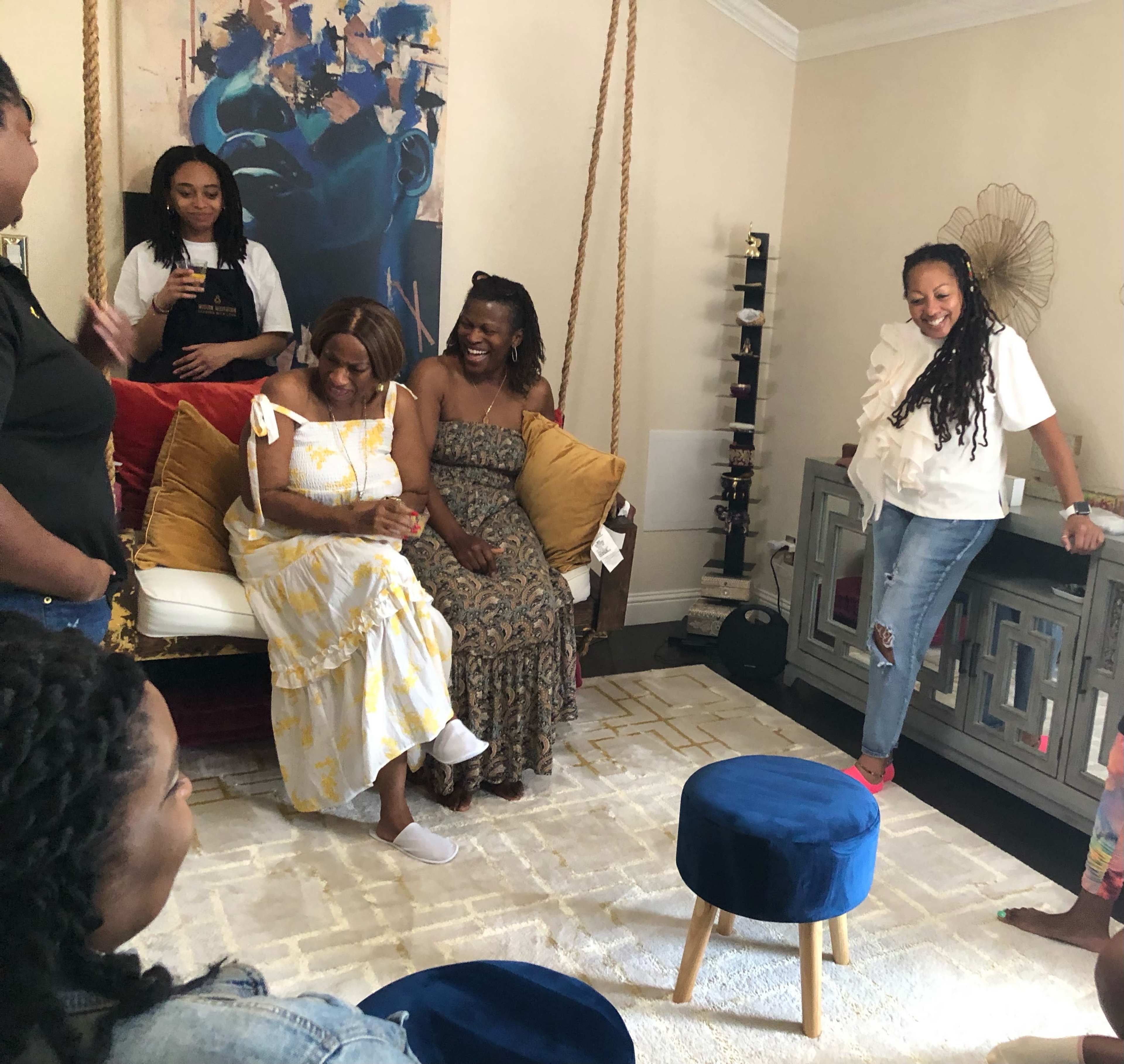 A group of six women gather in a living room, sitting and standing around a couch adorned with decorative pillows and a rug, engaging in conversation.