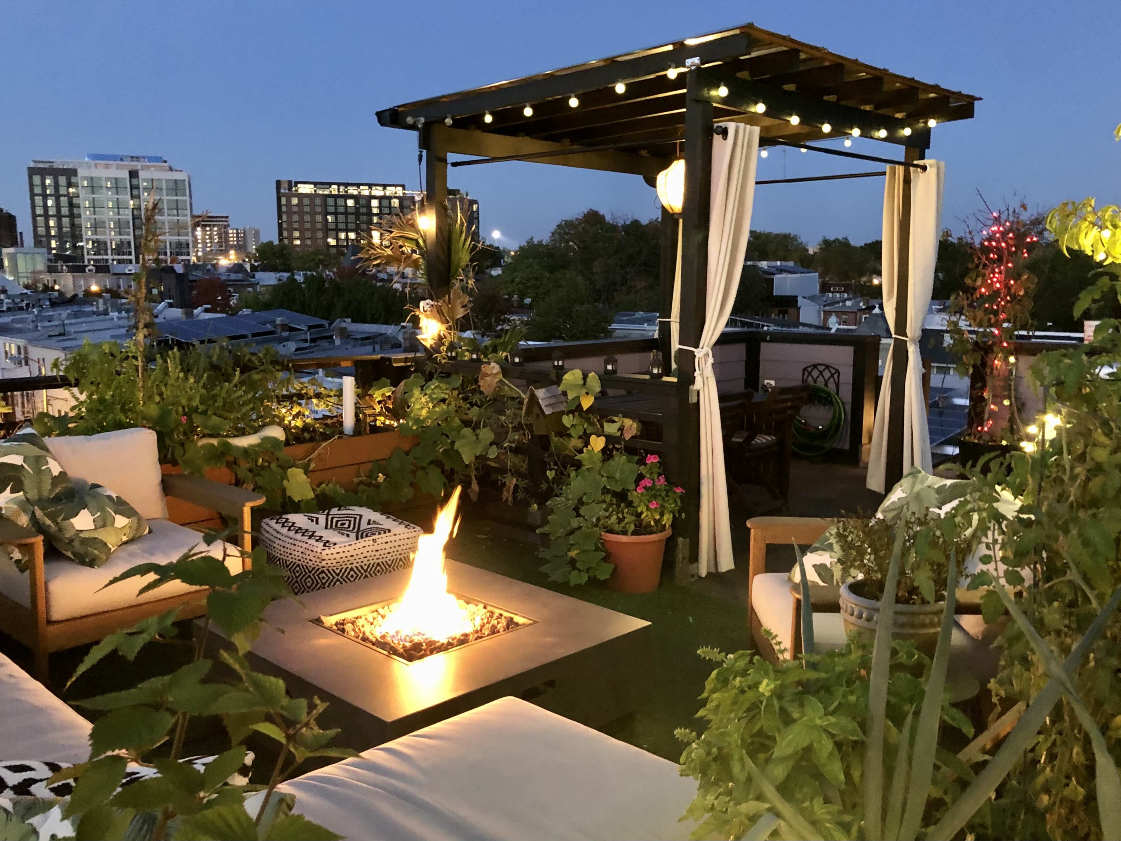 A rooftop terrace adorned with plants, comfortable seating, and a fire pit, illuminated by string lights at dusk.