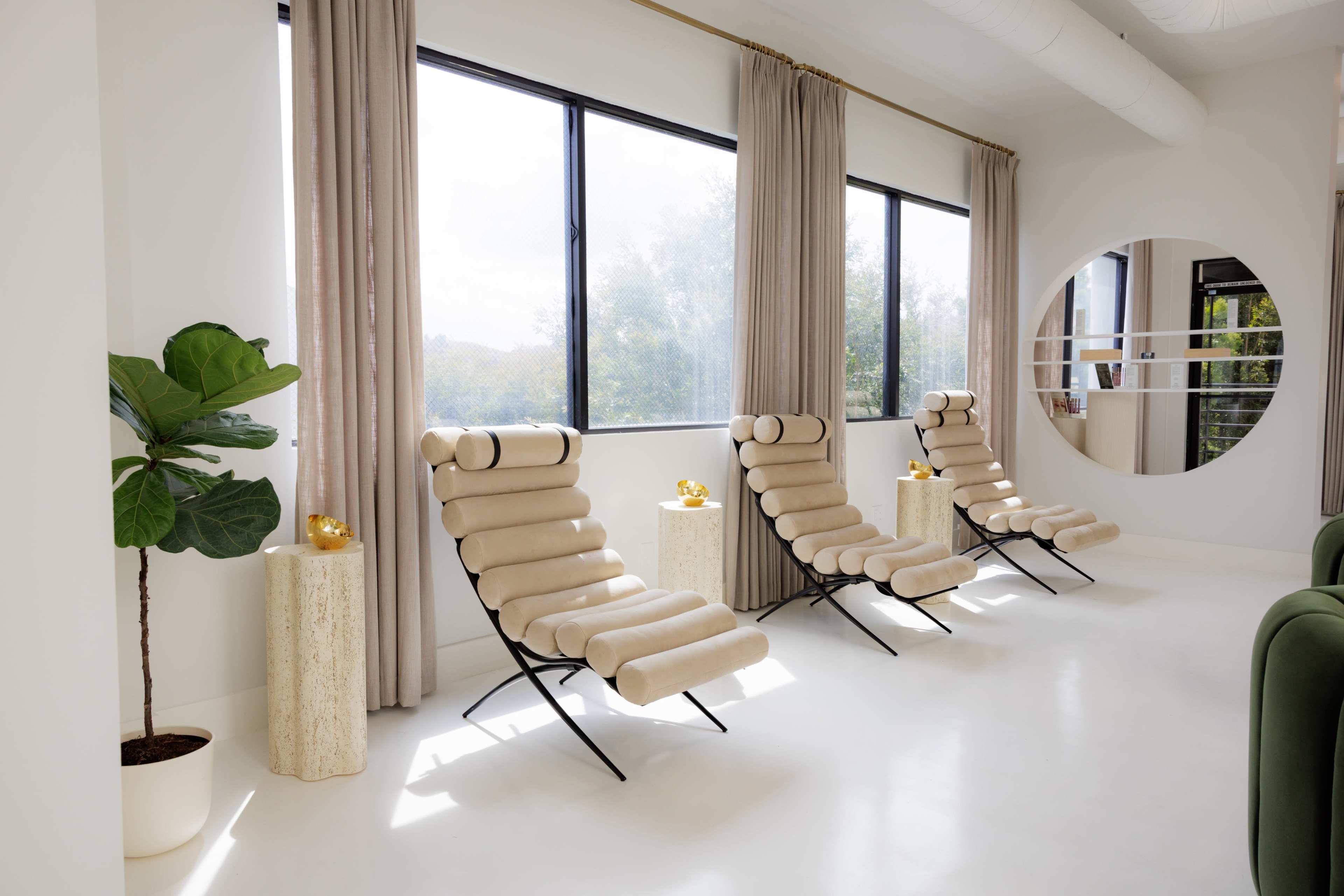 The image shows a bright and modern room with three beige lounge chairs aligned by large windows, complemented by a potted plant and decorative objects on white pedestals.