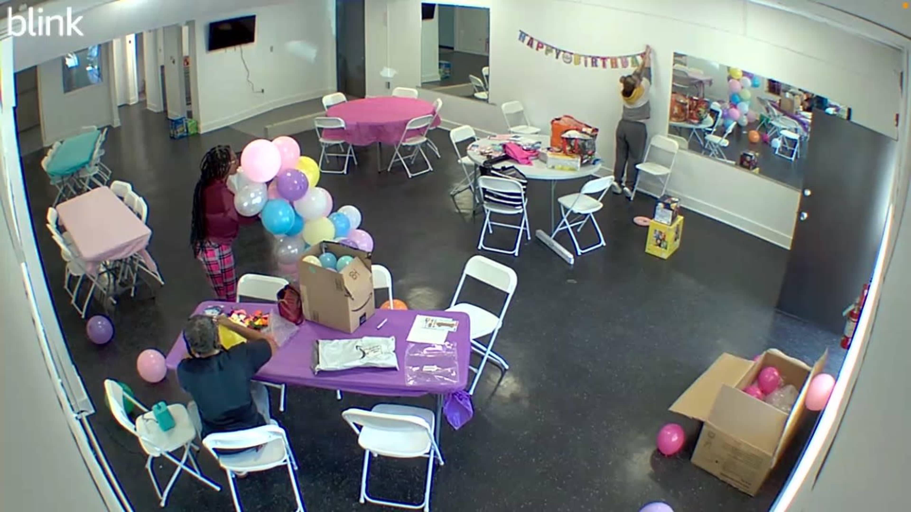 A party decor setup is taking place in a room with tables covered in pink and purple cloths, as one person hangs a banner while another arranges balloons.