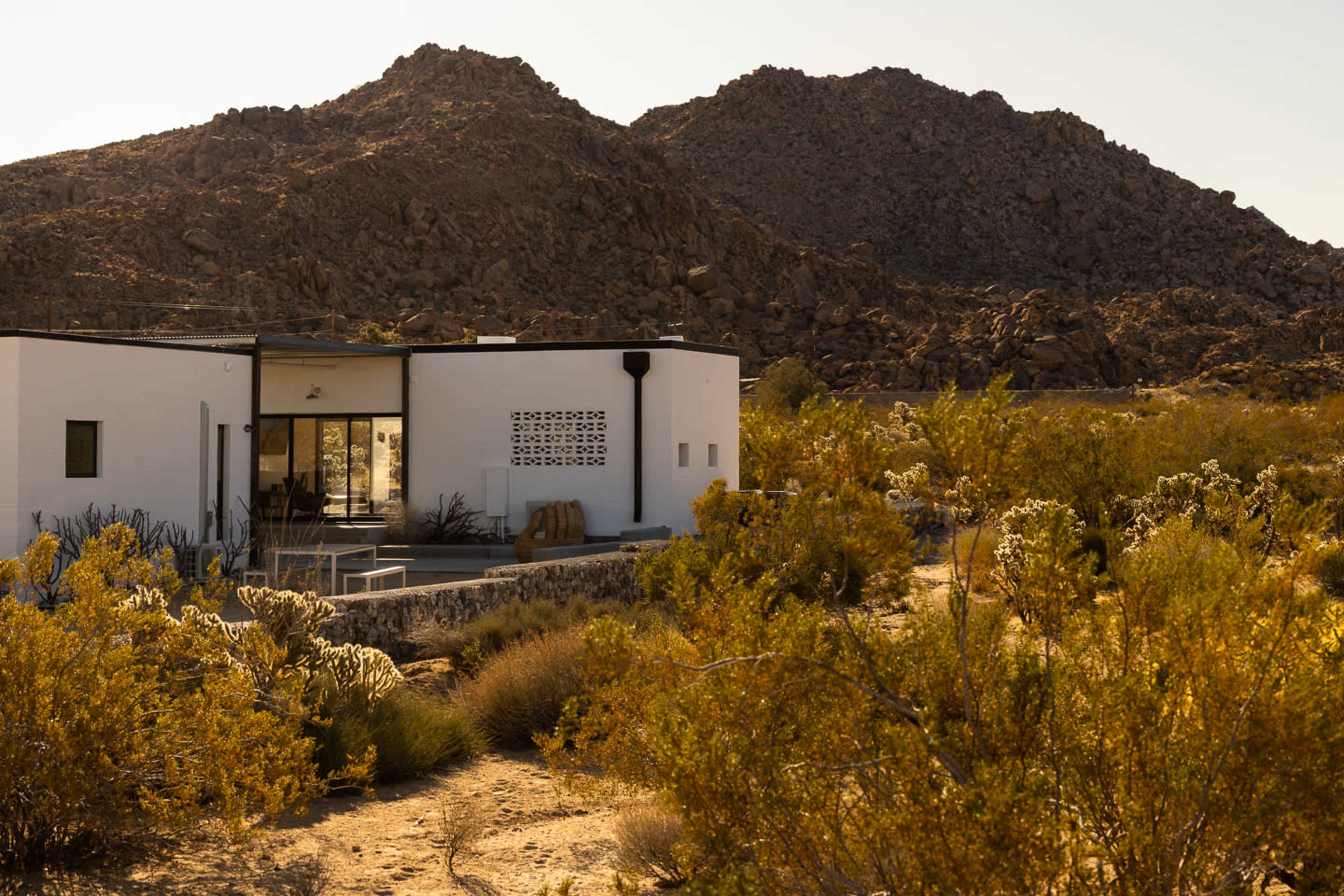 A modern white house sits against a backdrop of rocky mountains, surrounded by desert vegetation.