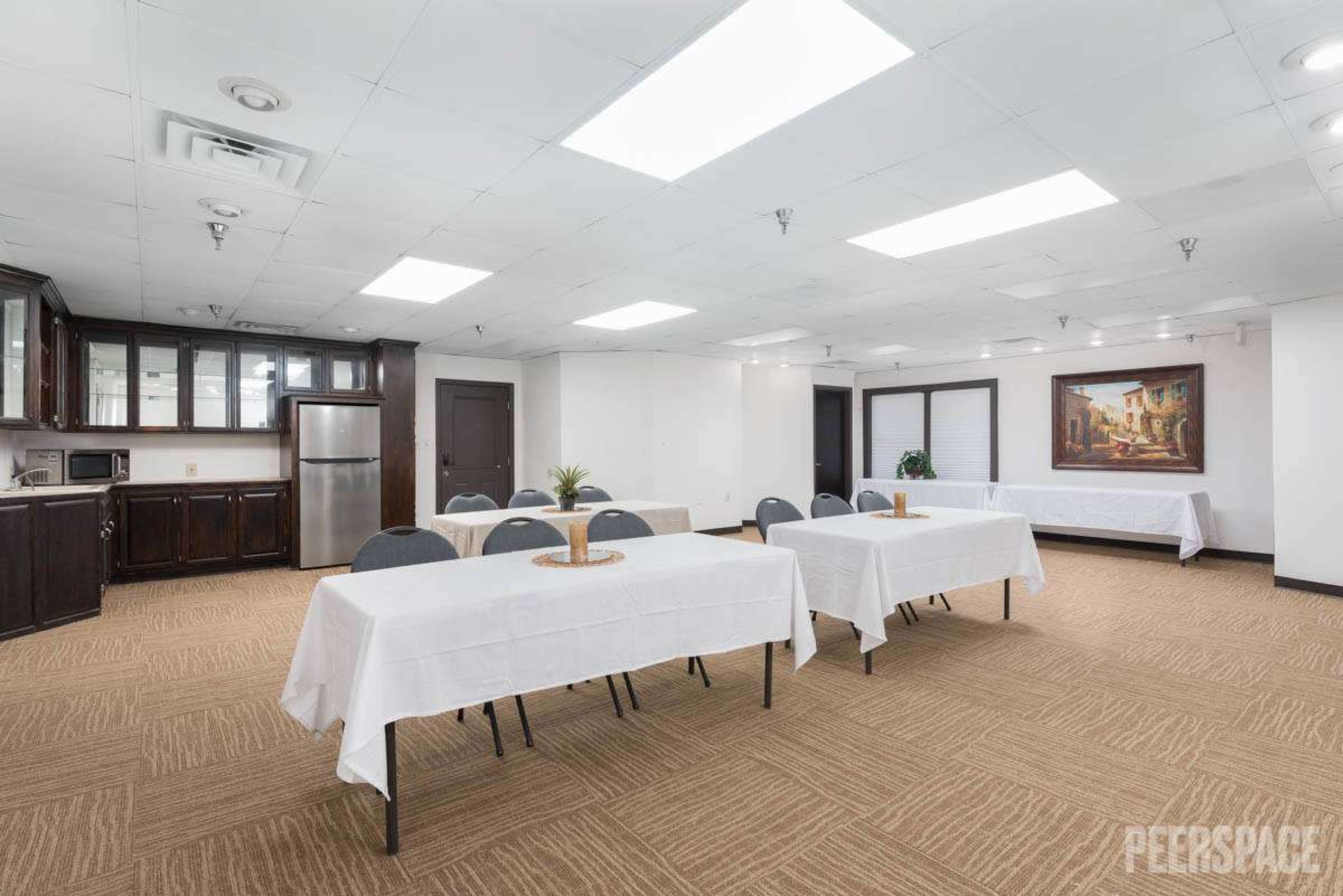 A conference room with several tables covered in white tablecloths, equipped with a kitchenette and a large painting on the wall.