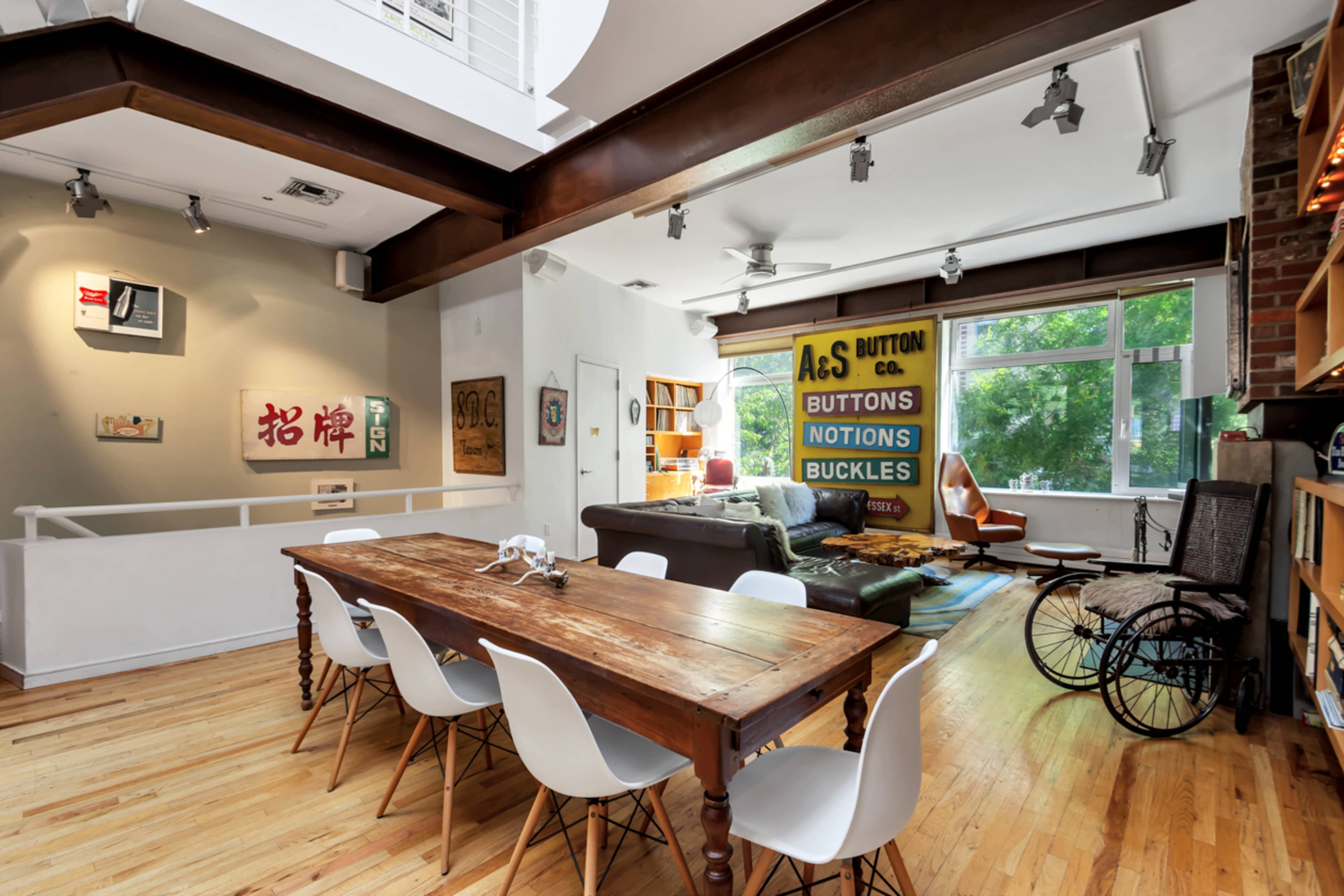 A spacious living area with a wooden dining table, white chairs, a leather sofa, and large windows allowing natural light to enter.