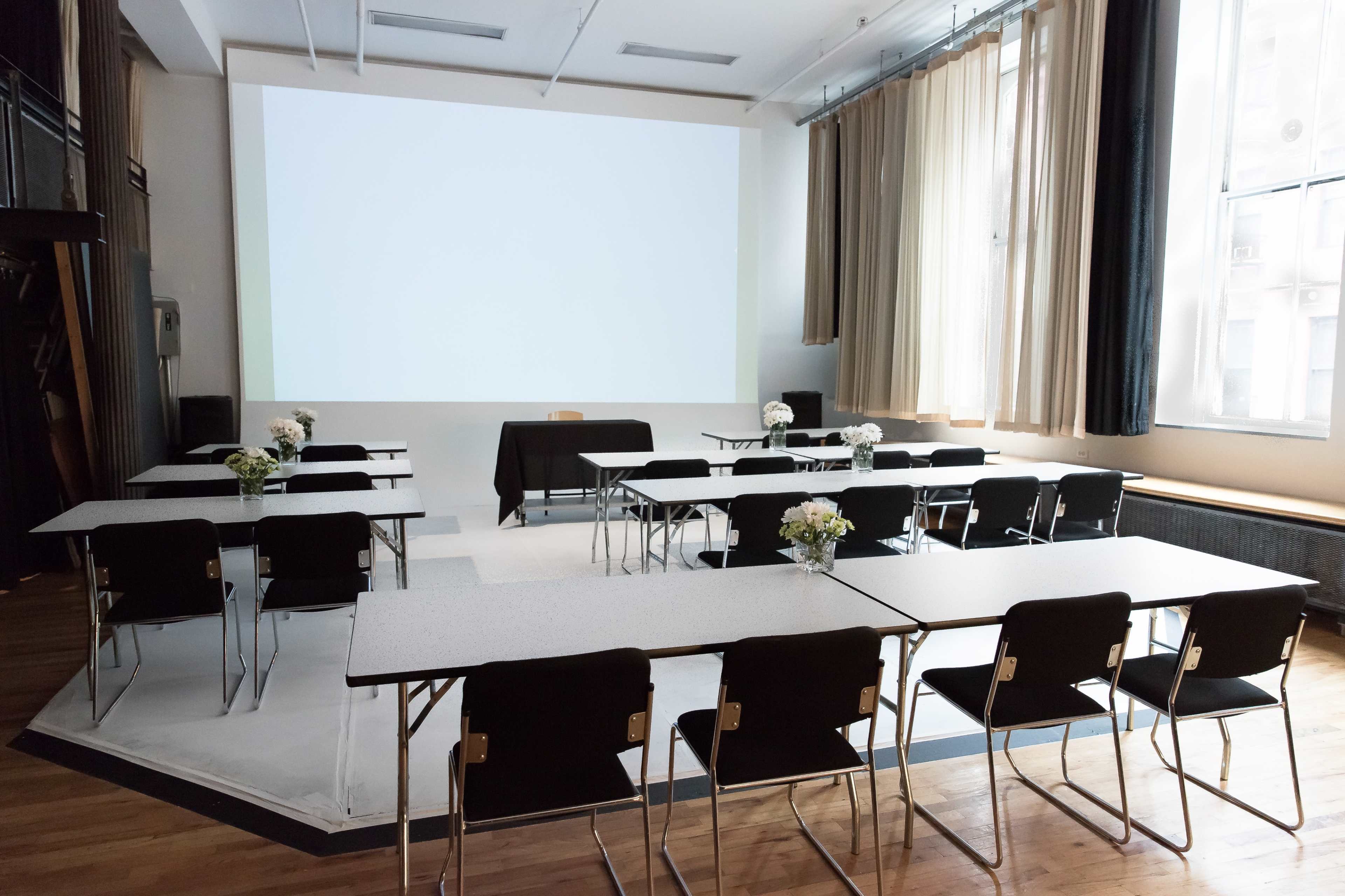 A seminar room with multiple tables and chairs arranged in a classroom setup, featuring a blank screen at the front and a table with a black cloth.