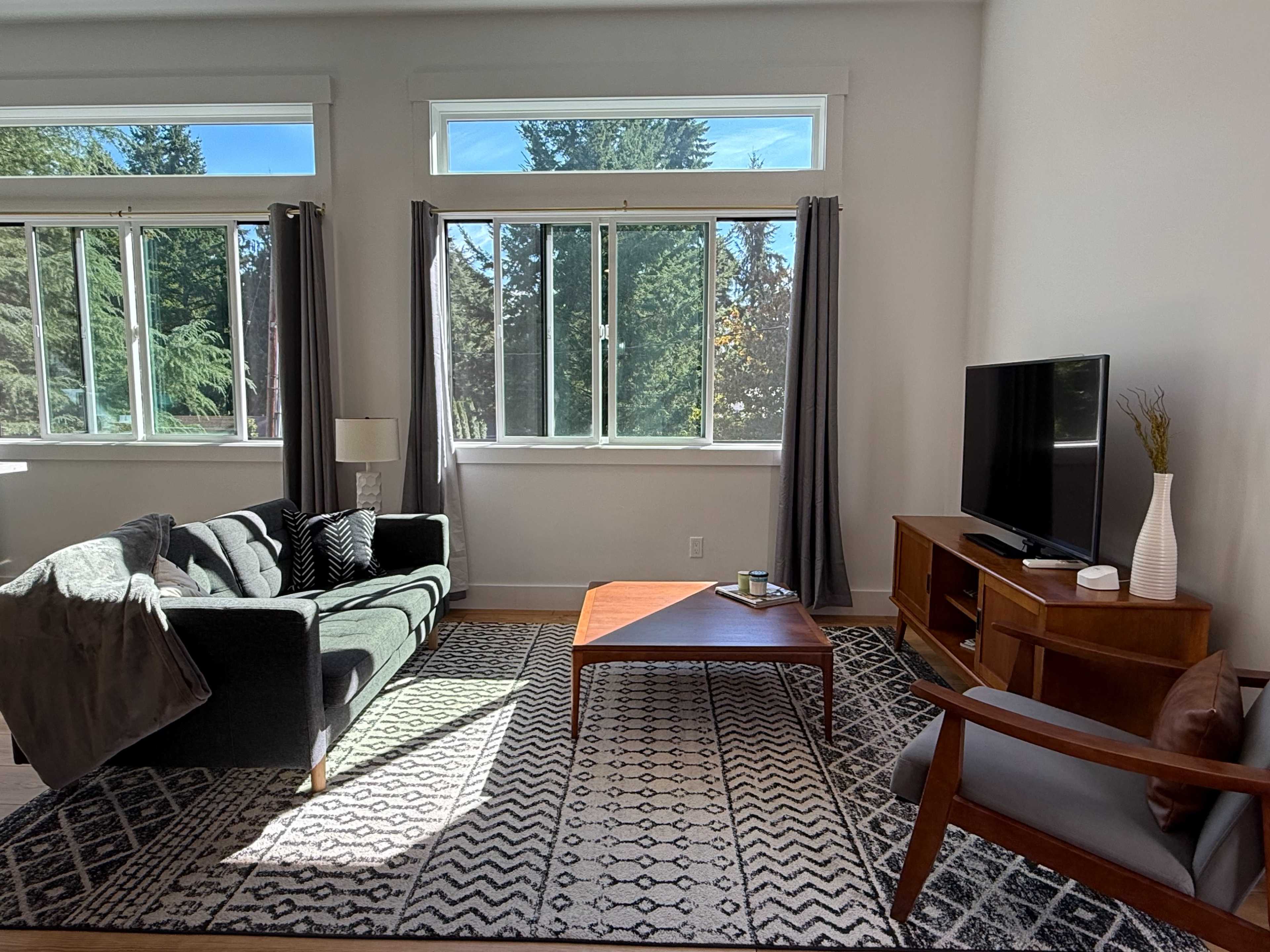 A modern living room features a dark sofa, a wooden coffee table, and a television in front of large windows with trees visible outside.