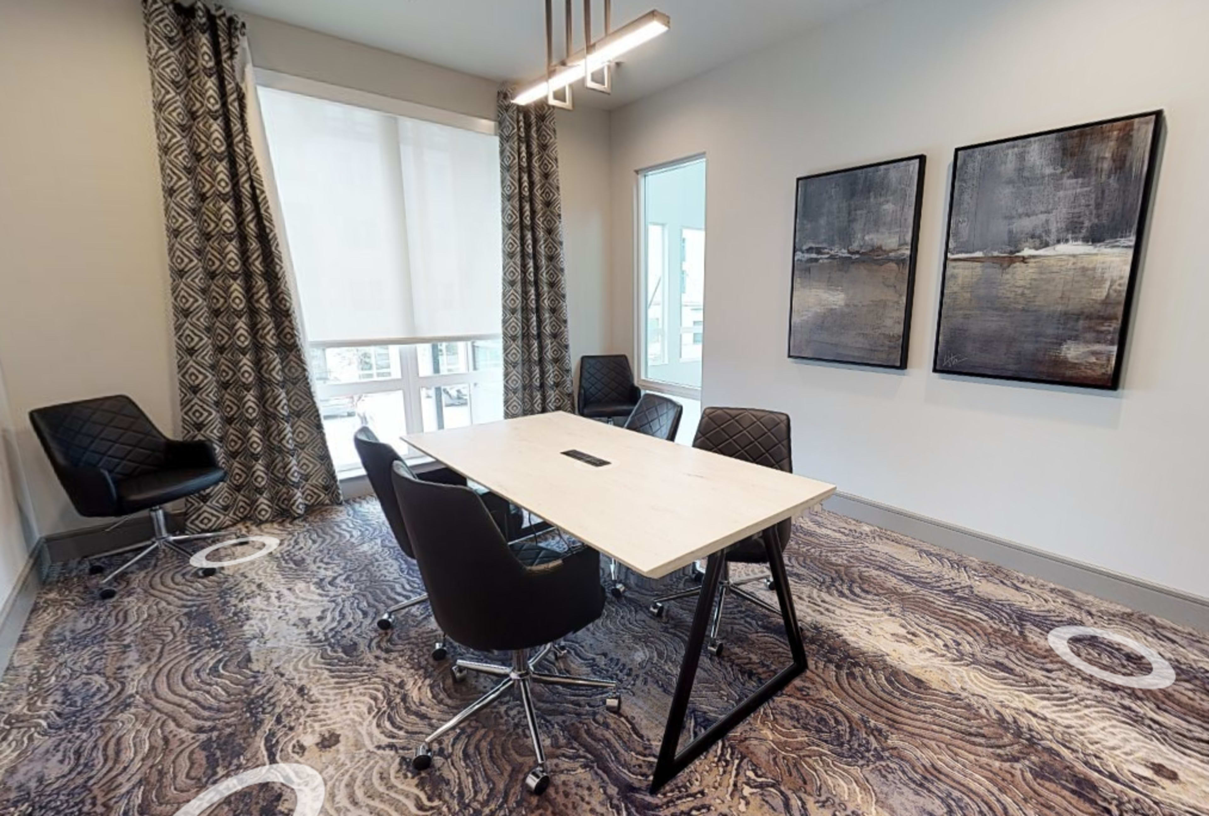 A small conference room features a rectangular table surrounded by four black chairs, with two abstract paintings on the wall and large windows covered by light-colored curtains.