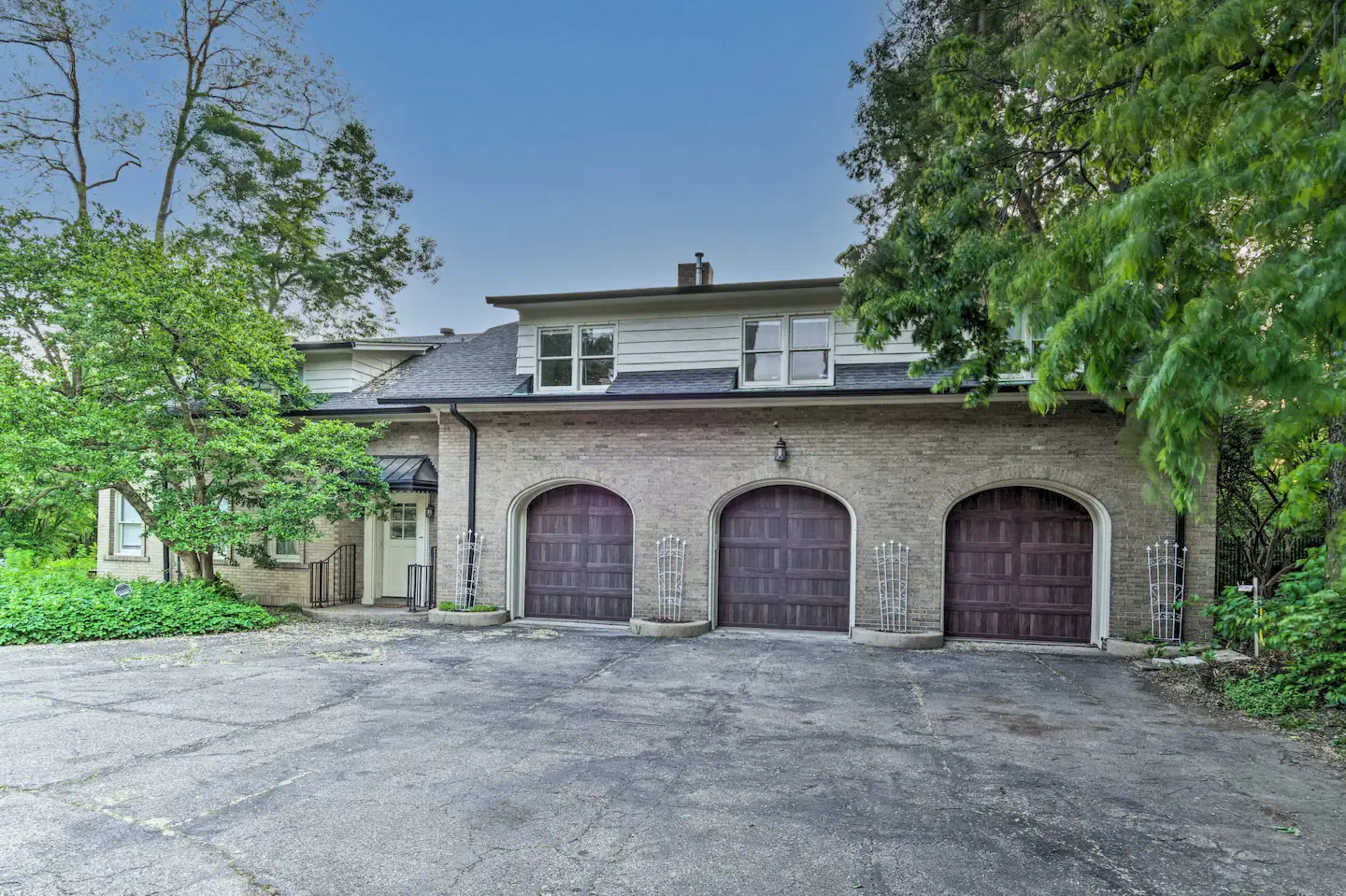 The image shows a large brick house with three garage doors, surrounded by trees and a paved driveway.