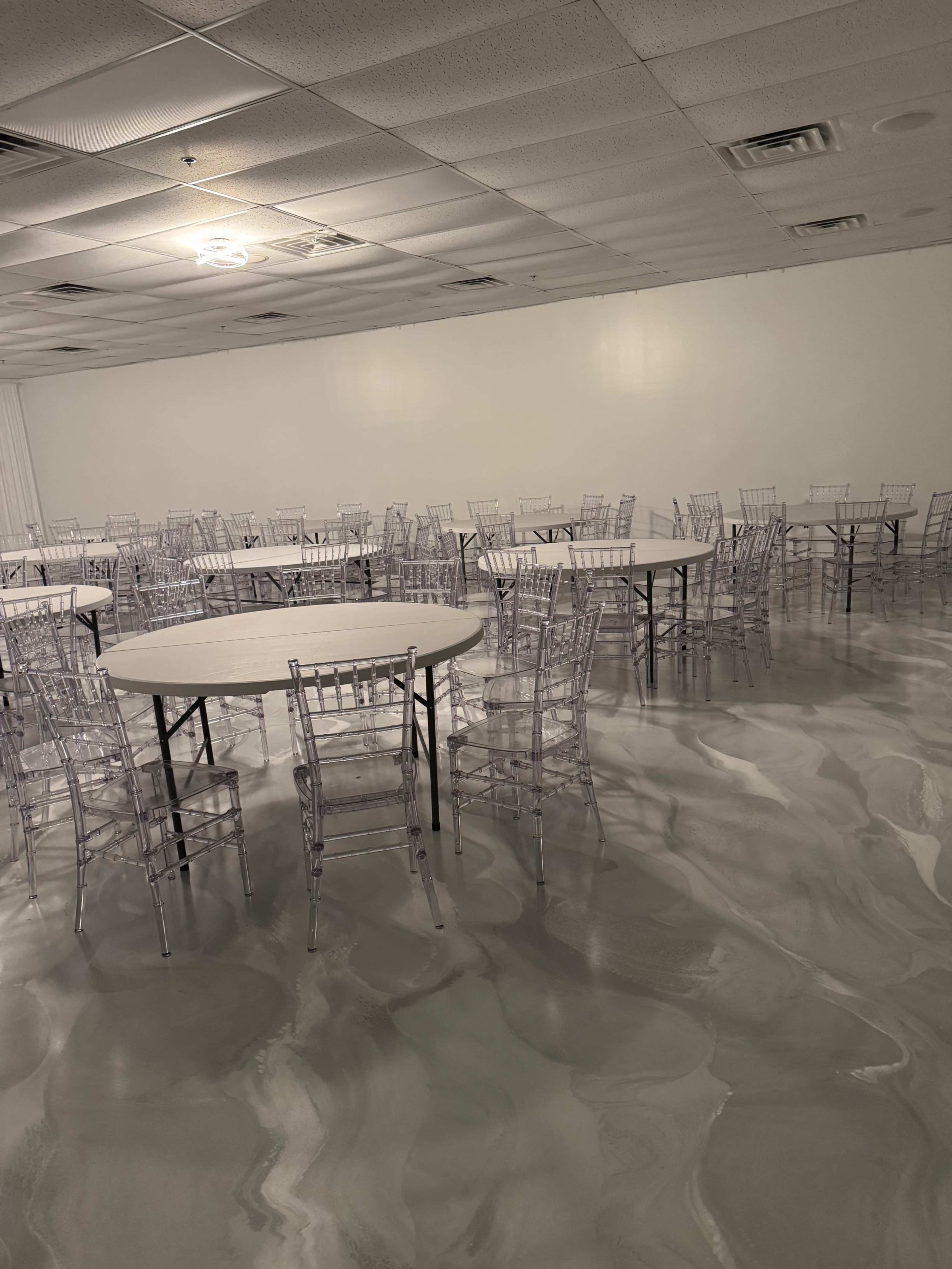 The image shows a large, empty event space with multiple round tables surrounded by clear chiavari chairs, set on a marbled floor.