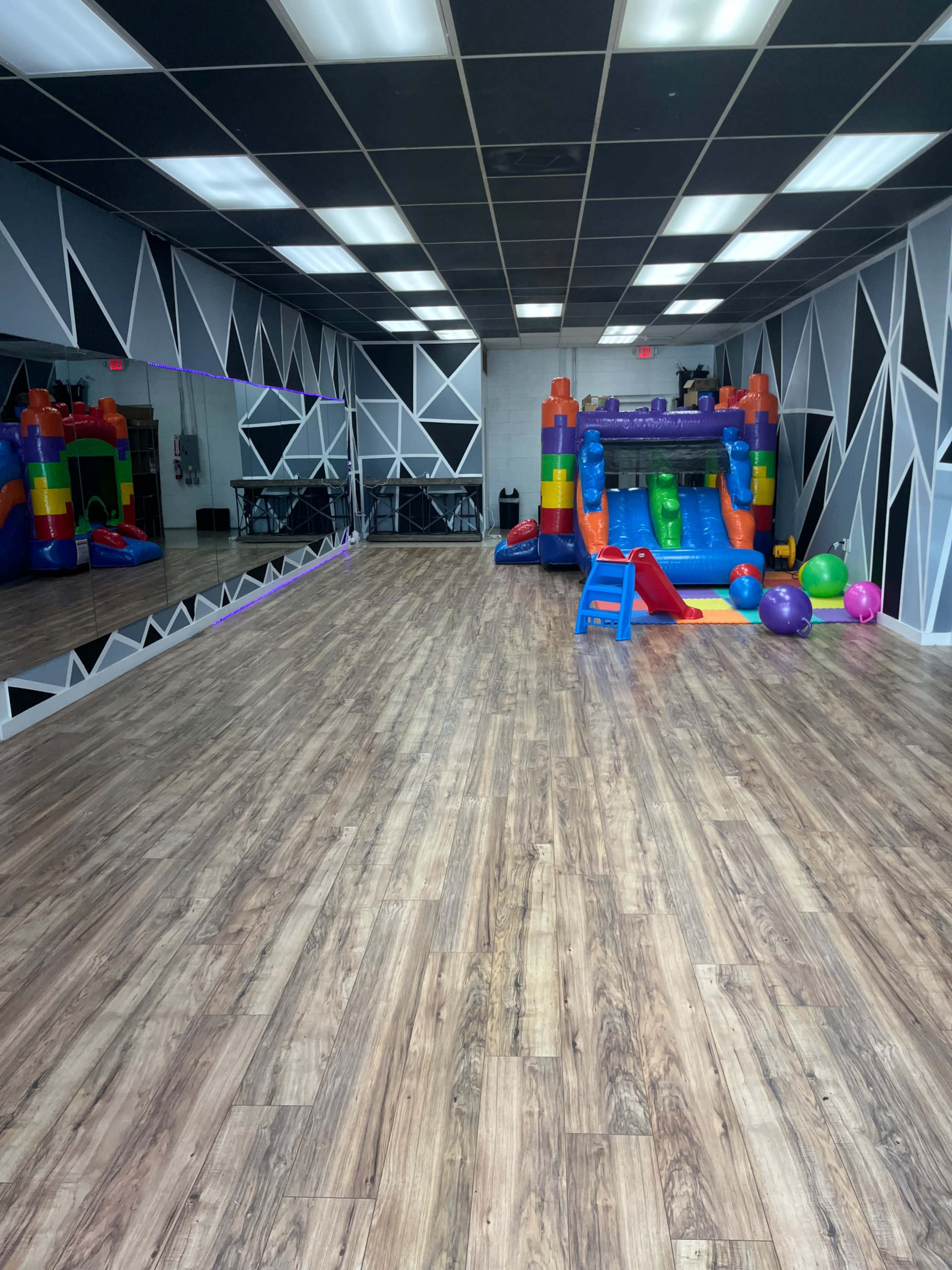 A spacious room with wood-style flooring, featuring a colorful inflatable bounce house and various play equipment at one end.