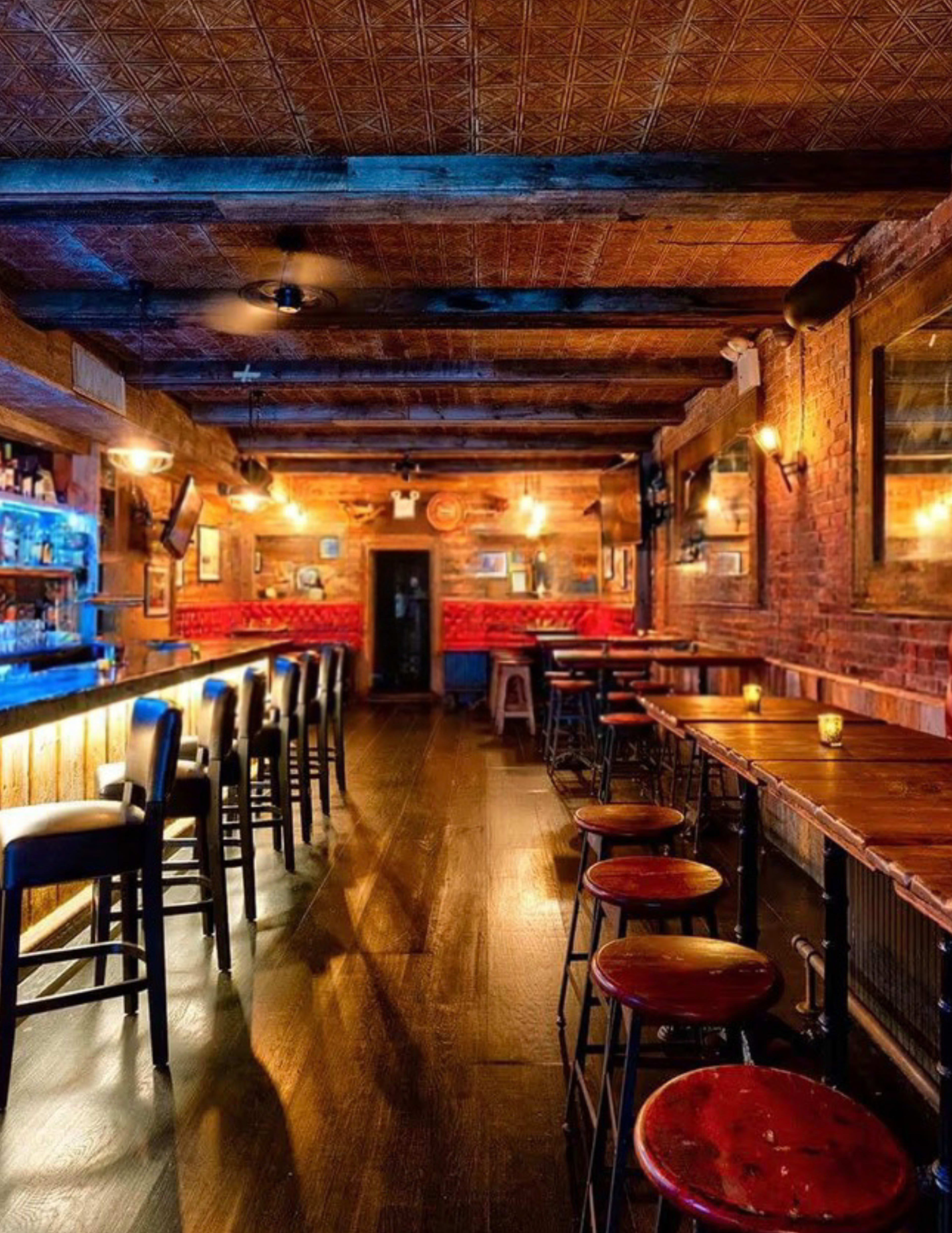The image shows an interior of a rustic bar with wooden floors, a long counter with bar stools, and warm lighting along the walls.