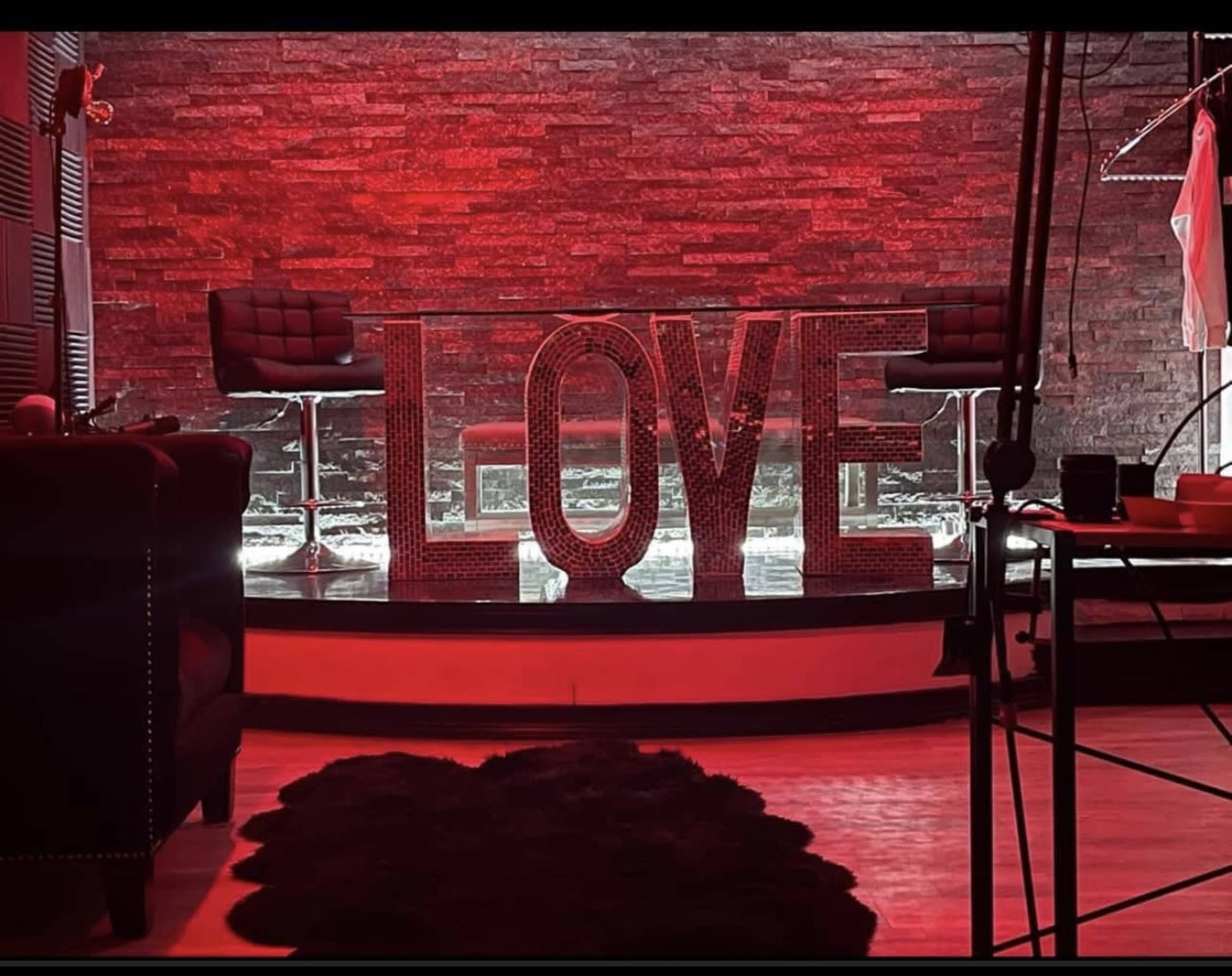 The image features a red-lit interior with large, illuminated letters spelling "LOVE" on a stage-like setup, surrounded by modern chairs and a decorative rug.