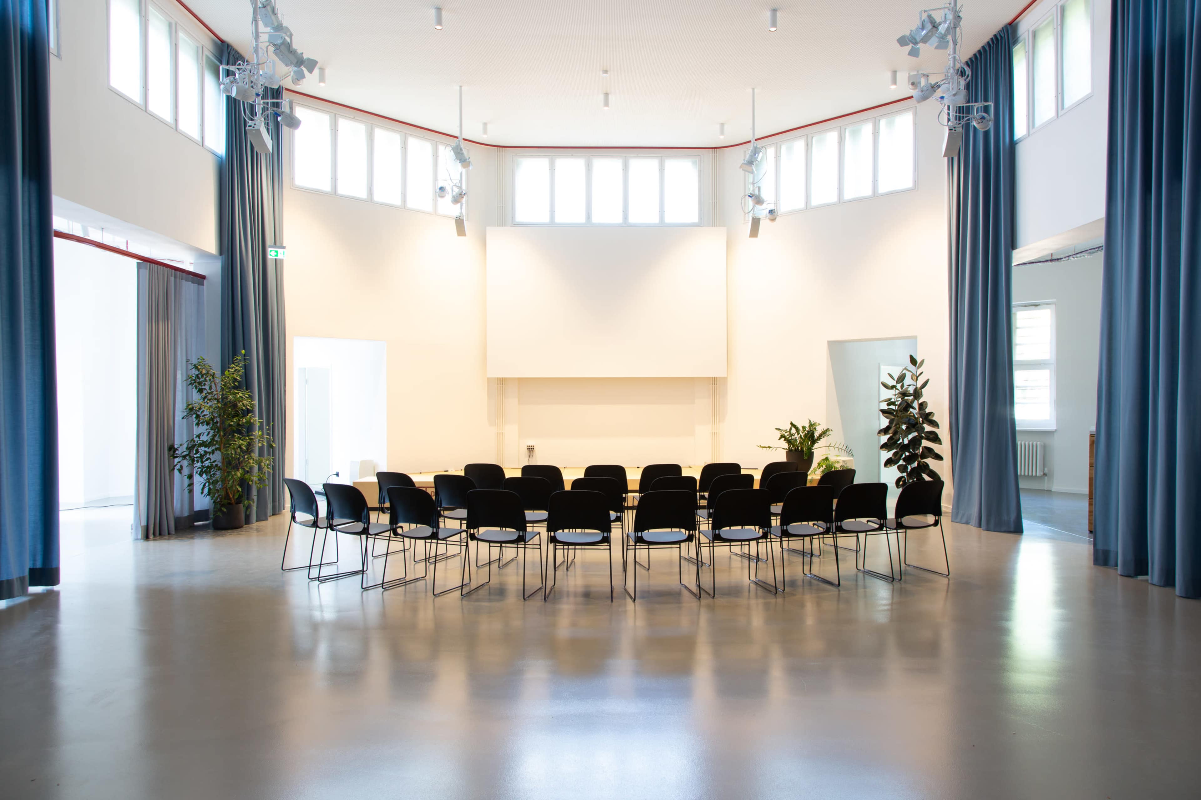 A row of black chairs is arranged in a semi-circle in a bright, minimalist room with large windows and green plants.