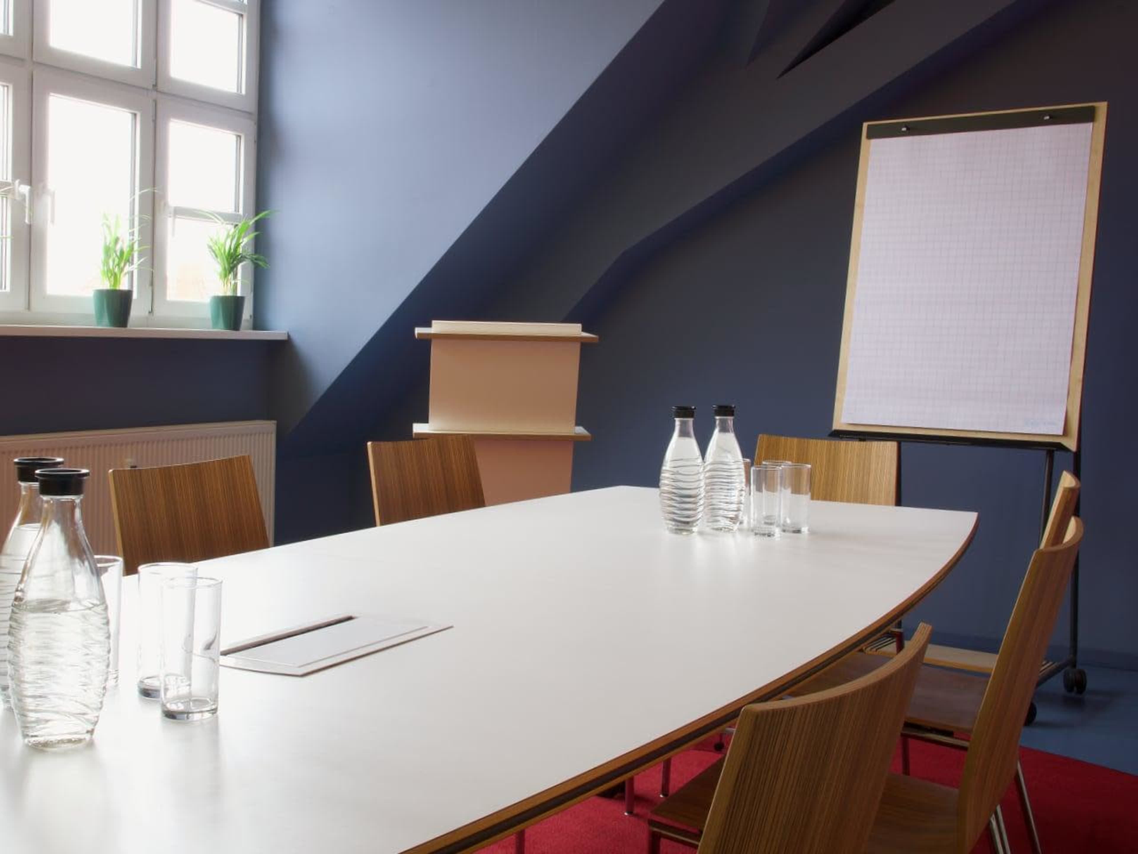 A conference room features a long white table with water bottles and glasses, a blank flip chart, and large windows with potted plants.
