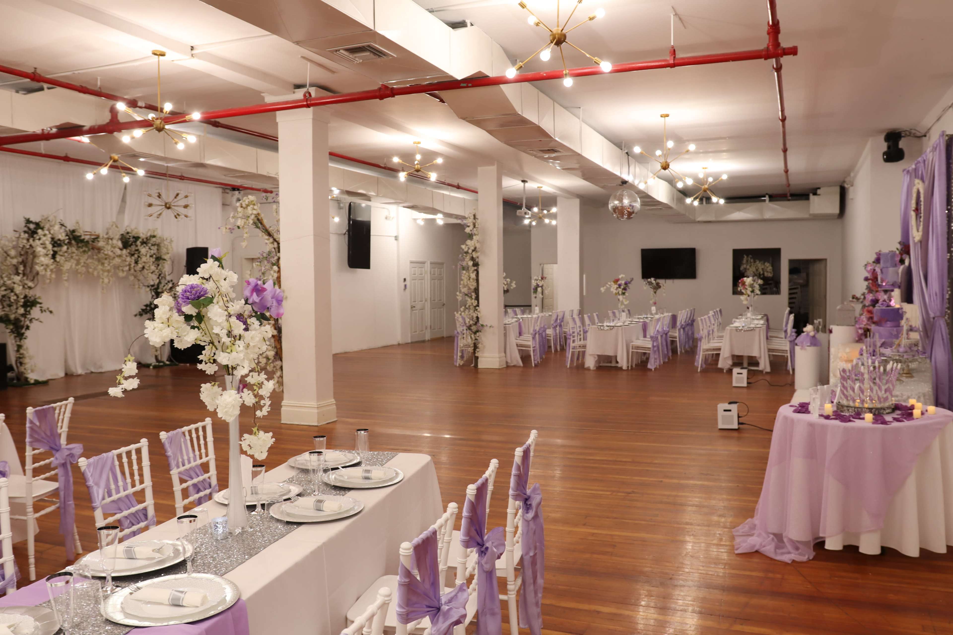 A spacious event hall is set up for a formal gathering, featuring tables adorned with purple decorations and floral arrangements.