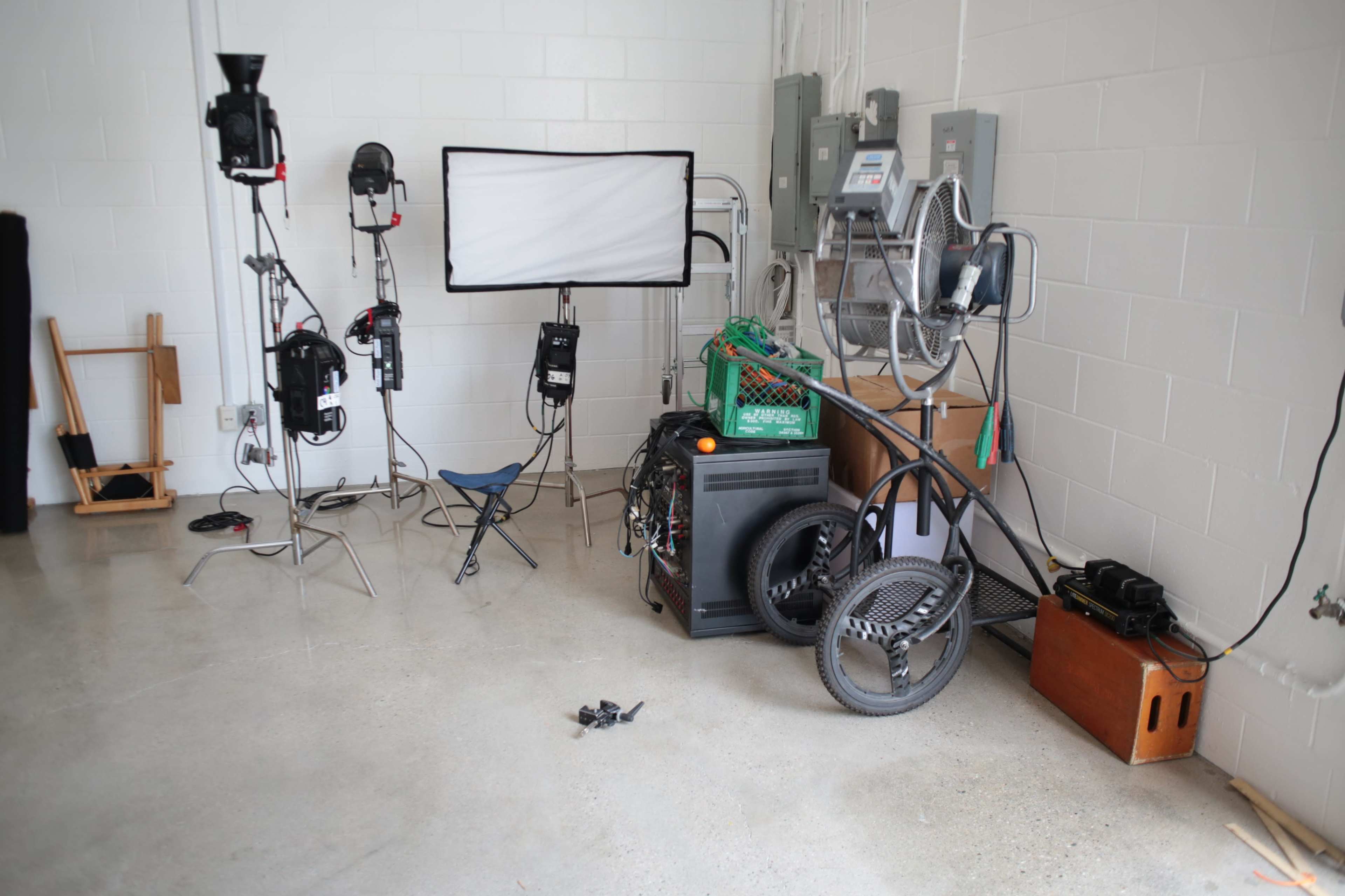 A cluttered corner of a studio with lighting equipment, a folding chair, and various objects including a cart and old machinery against a white wall.
