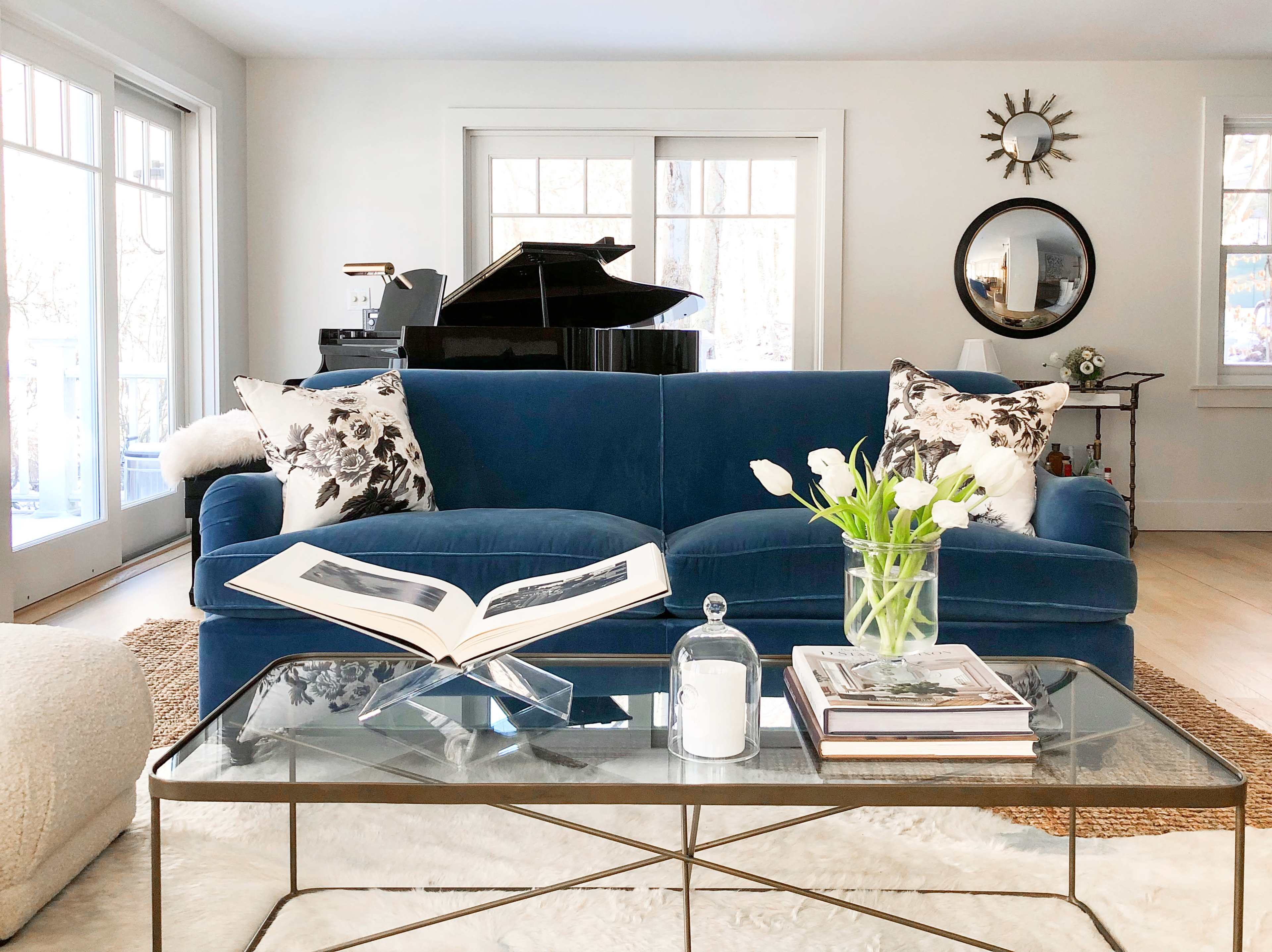 A blue sofa with floral-patterned pillows is positioned in a bright room, featuring a glass coffee table and a grand piano in the background.
